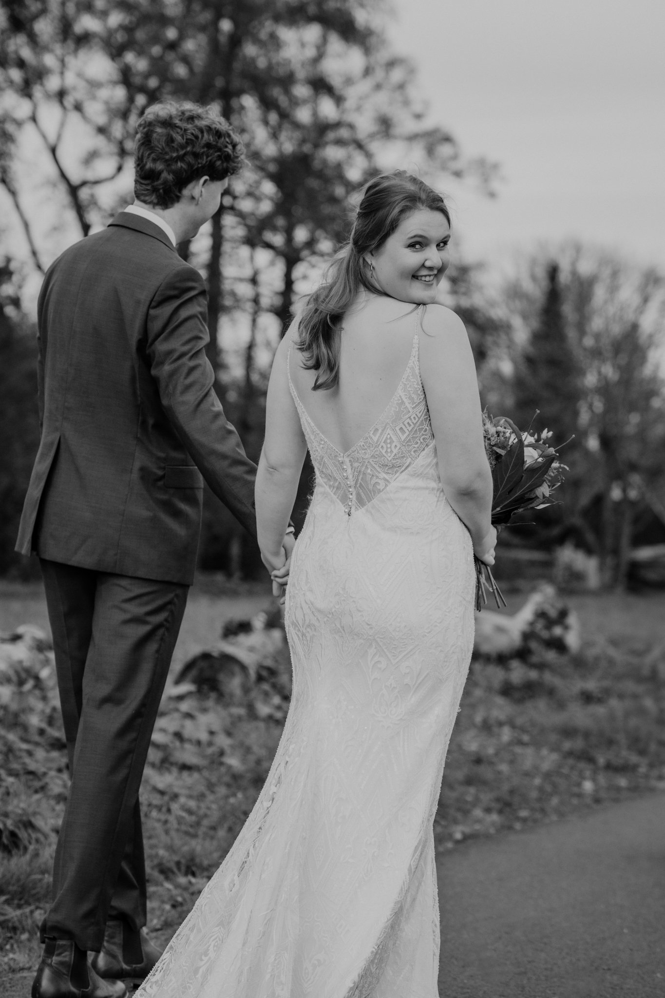 Een zwart-wit foto van een gelukkig bruidspaar dat hand in hand loopt, met de bruid die glimlacht naar de camera, in een park met bomen op de achtergrond.
