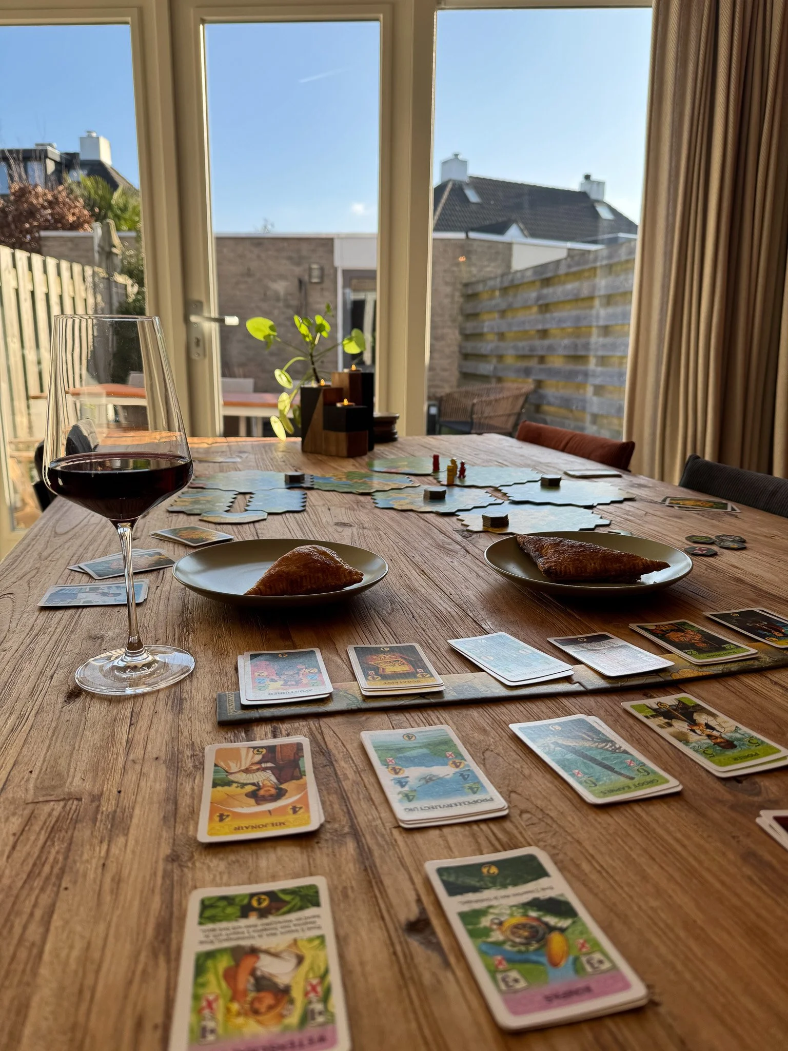 Een houten eettafel met een glas rode wijn, twee bordjes met een croissant, een aantal speelfiches en kaarten van een bordspel op een zonnige dag in een huis met grote ramen en een tuin.