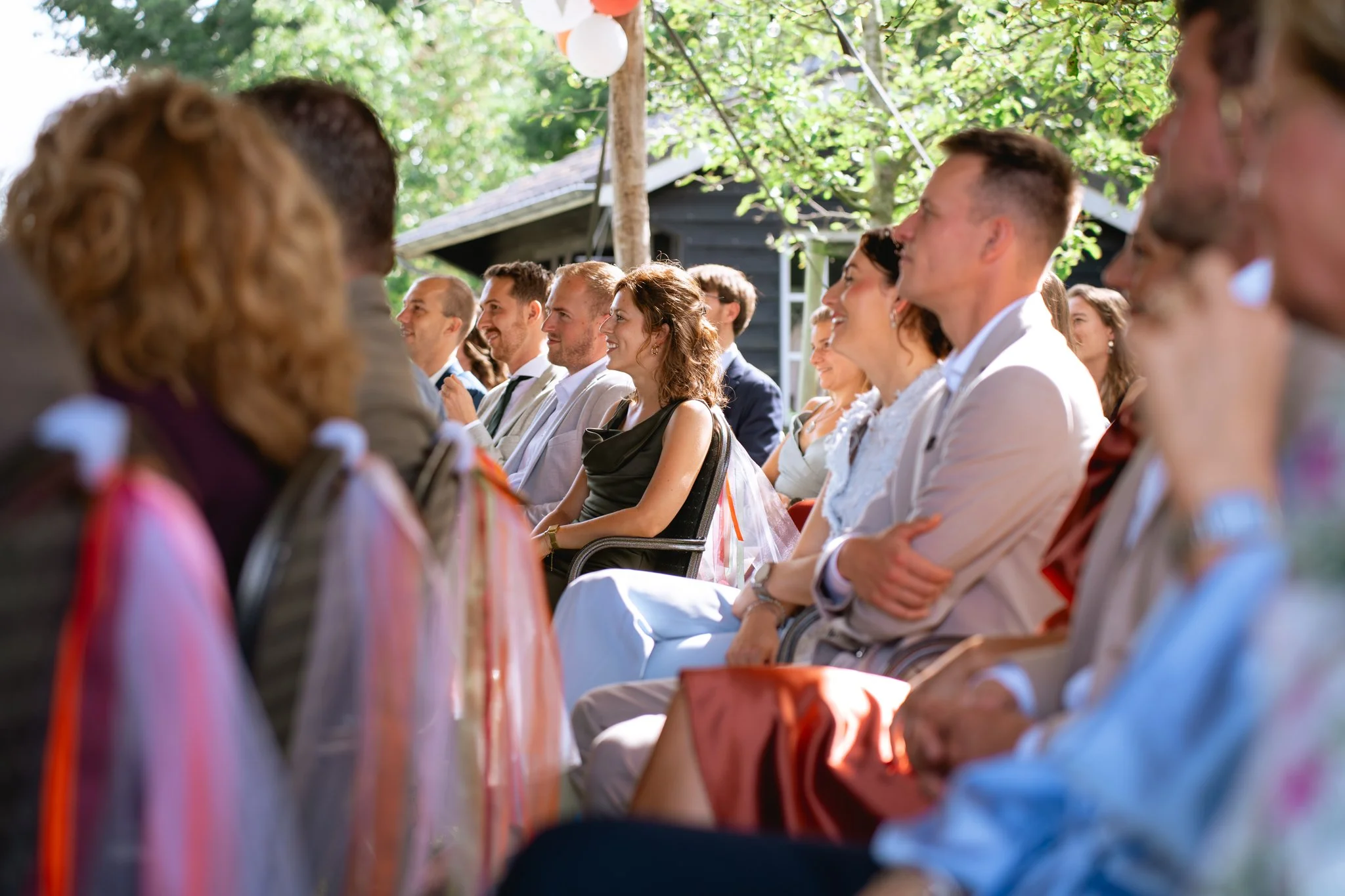 Gasten zitten aandachtig te kijken naar een huwelijsceremonie, buiten onder een tent