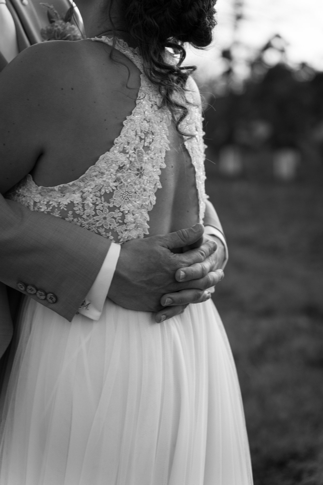 Een zwart-wit foto van een pasgetrouwd stel. De bruid draagt een jurk met kanten details en een open rug, terwijl de man in een pak staat. Ze omhelzen elkaar en houden elkaars handen vast.