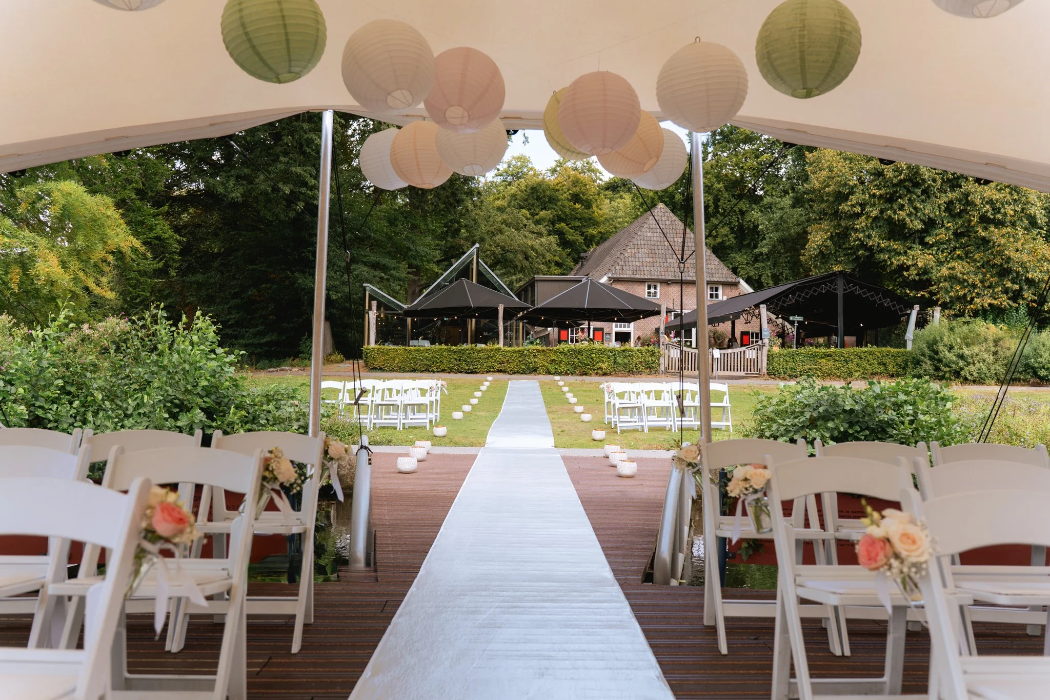 Een overdekte trouwlocatie met wit bloemenversiering en een witte strook die leidt naar een buitenruimte met zitplaatsen, gras en een huis op de achtergrond, versierd met papieren lampionnen.