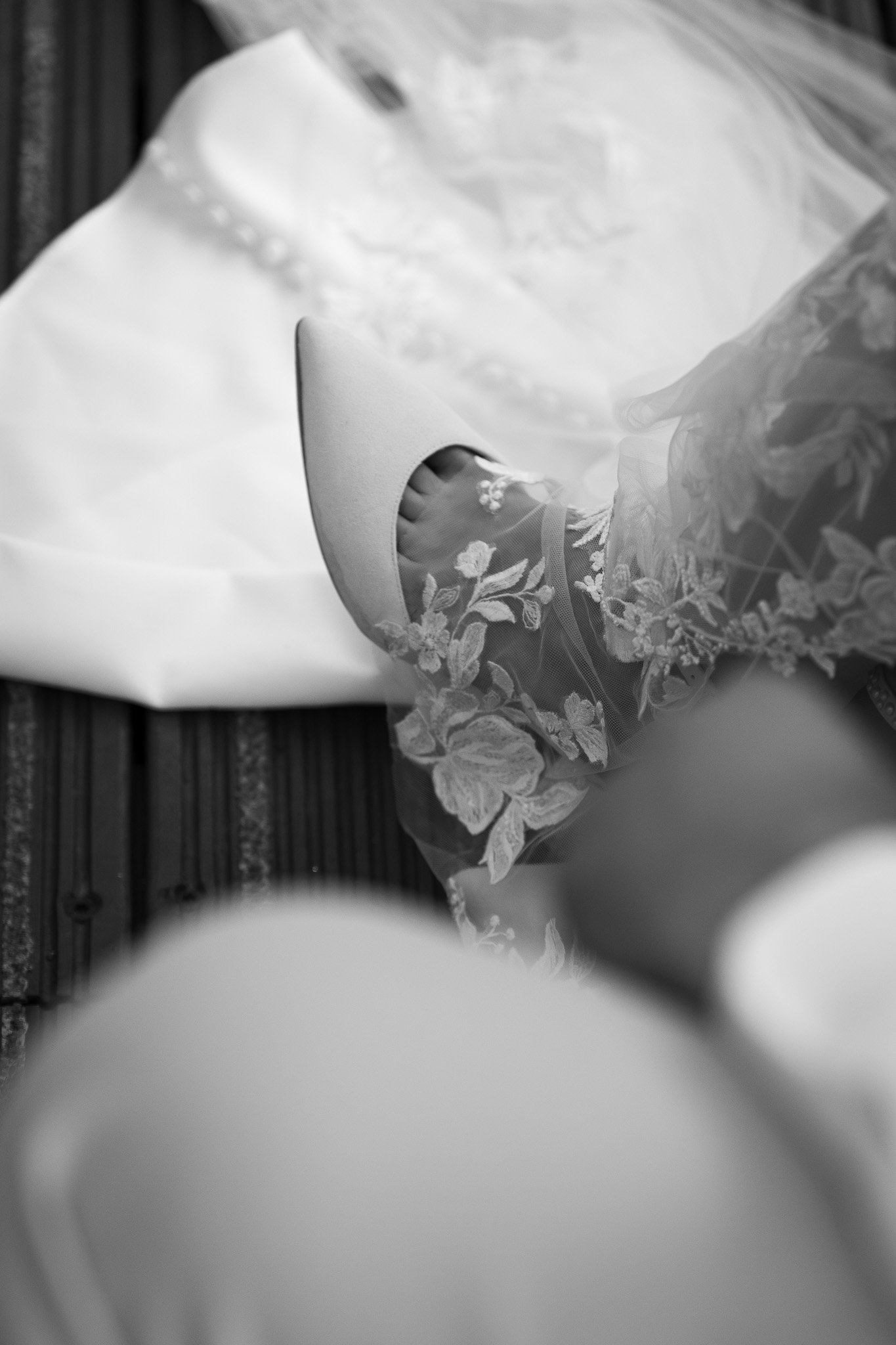 Close-up van een vrouwelijke voet in een hoge hak met decoratieve bloemen, gedragen op een houten vloer met een witte jurk ernaast.