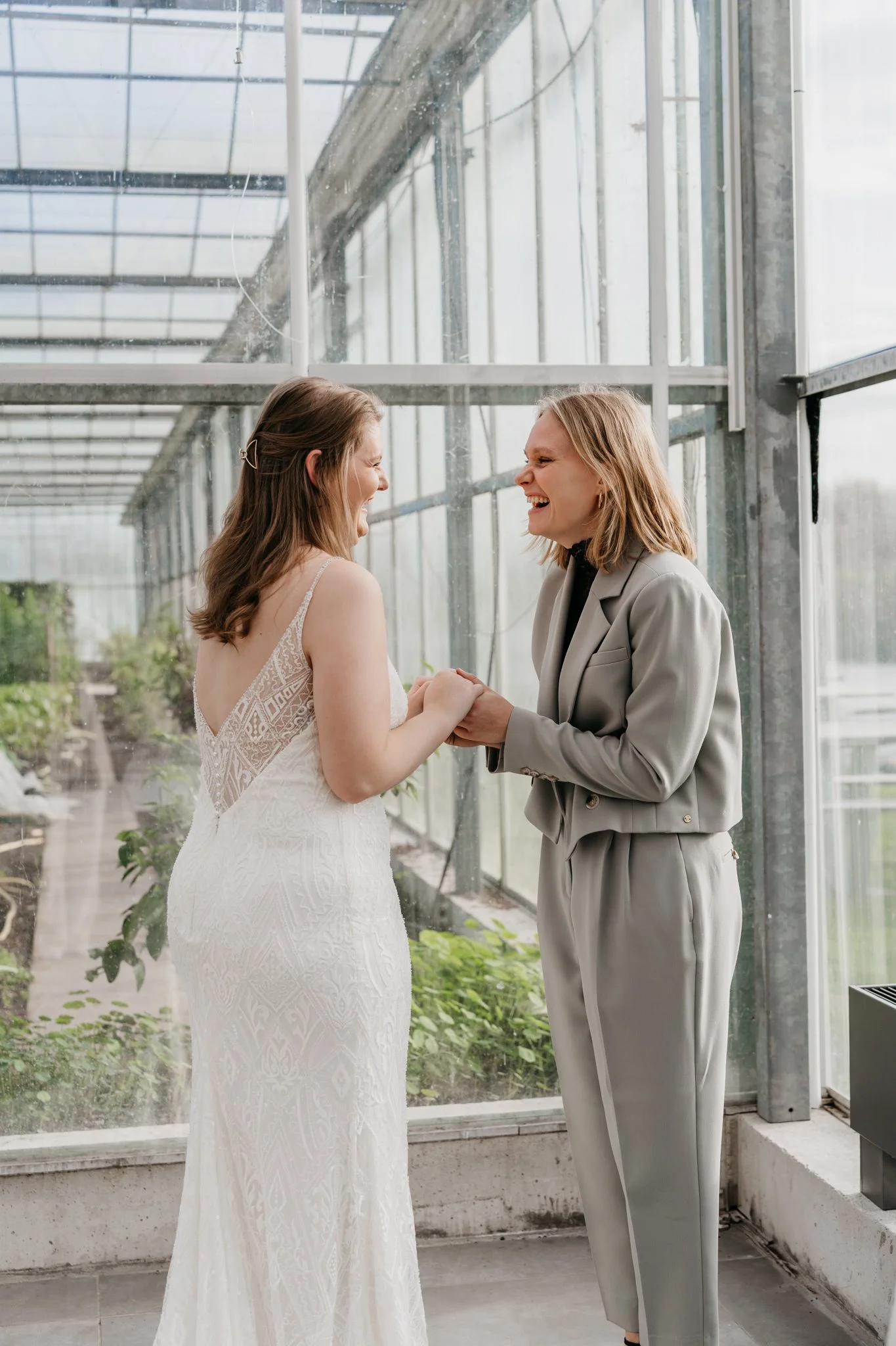 Twee vrouwen houden elkaars handen vast en lachen in een kas, een van hen draagt een bruidsjurk en de andere een lichtgrijze pak.