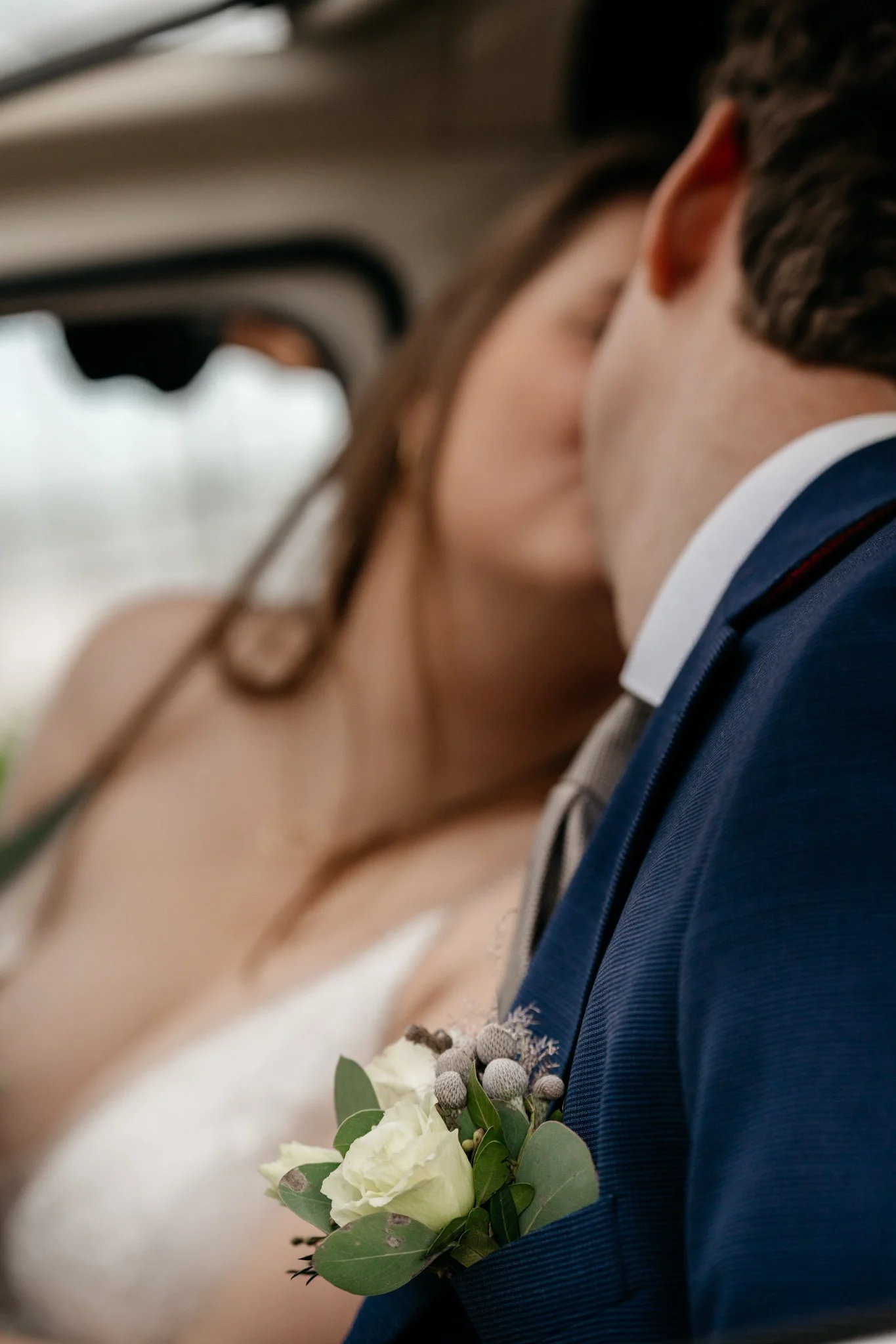 Close-up van een bruiloftskoppel dat elkaar kust, met een boutonnière van witte roos en groene bladeren op de bruidegom in een donkerblauw pak.