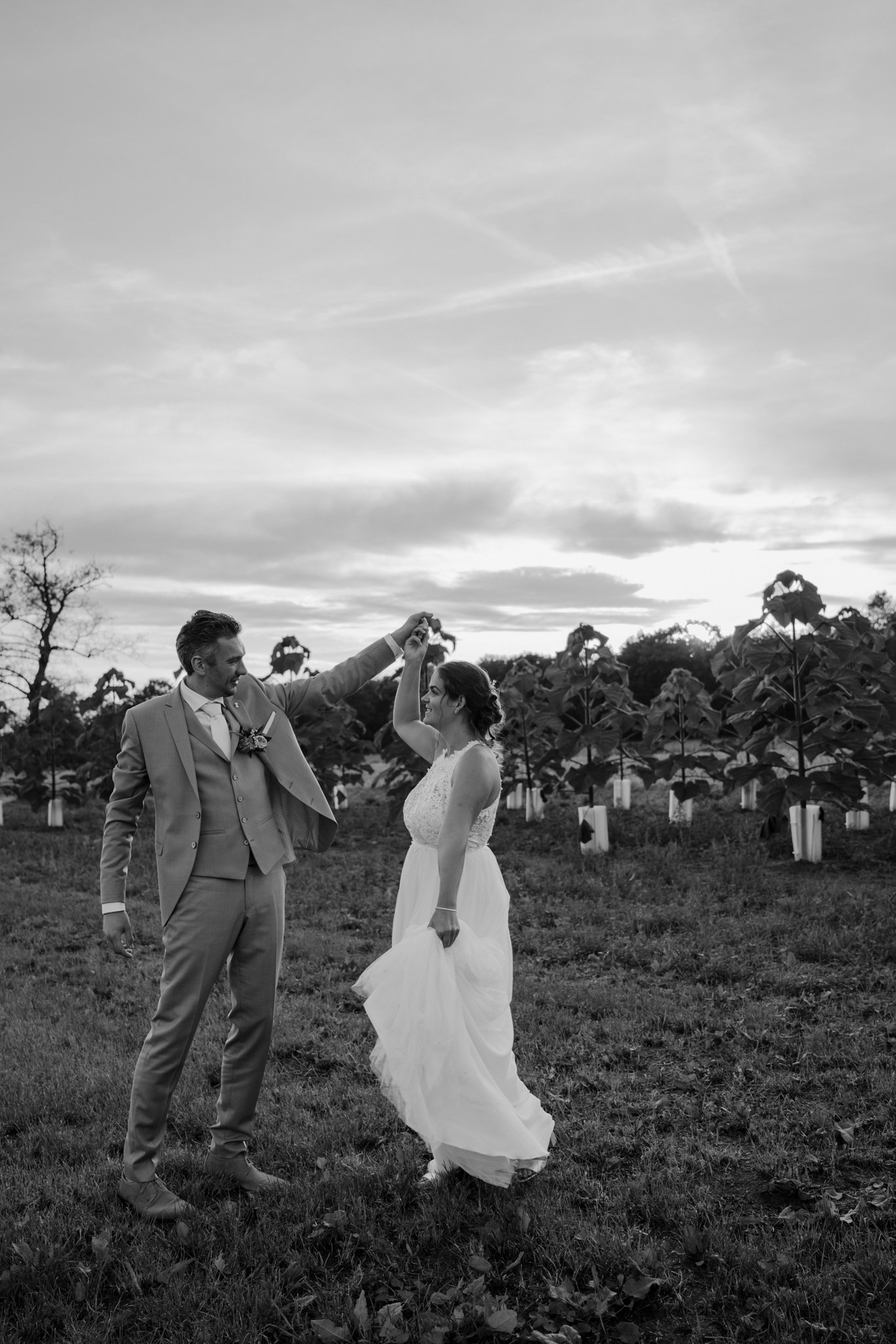 Een zwart-wit foto van een bruid en bruidegom die samen dansen in een open veld met bomen op de achtergrond, onder een bewolkte hemel.
