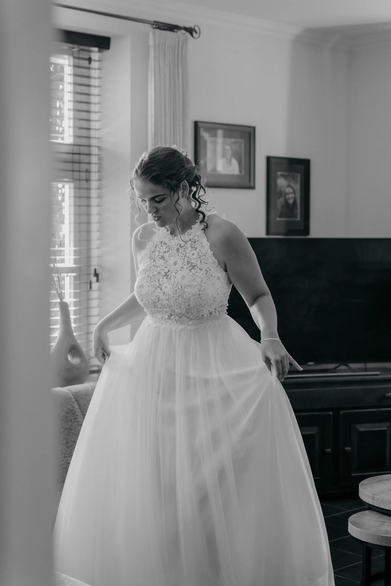 Vrouw in trouwjurk thuis aan het klaarmaken, in een interieur met foto's aan de muur en een venster met jaloezieën.