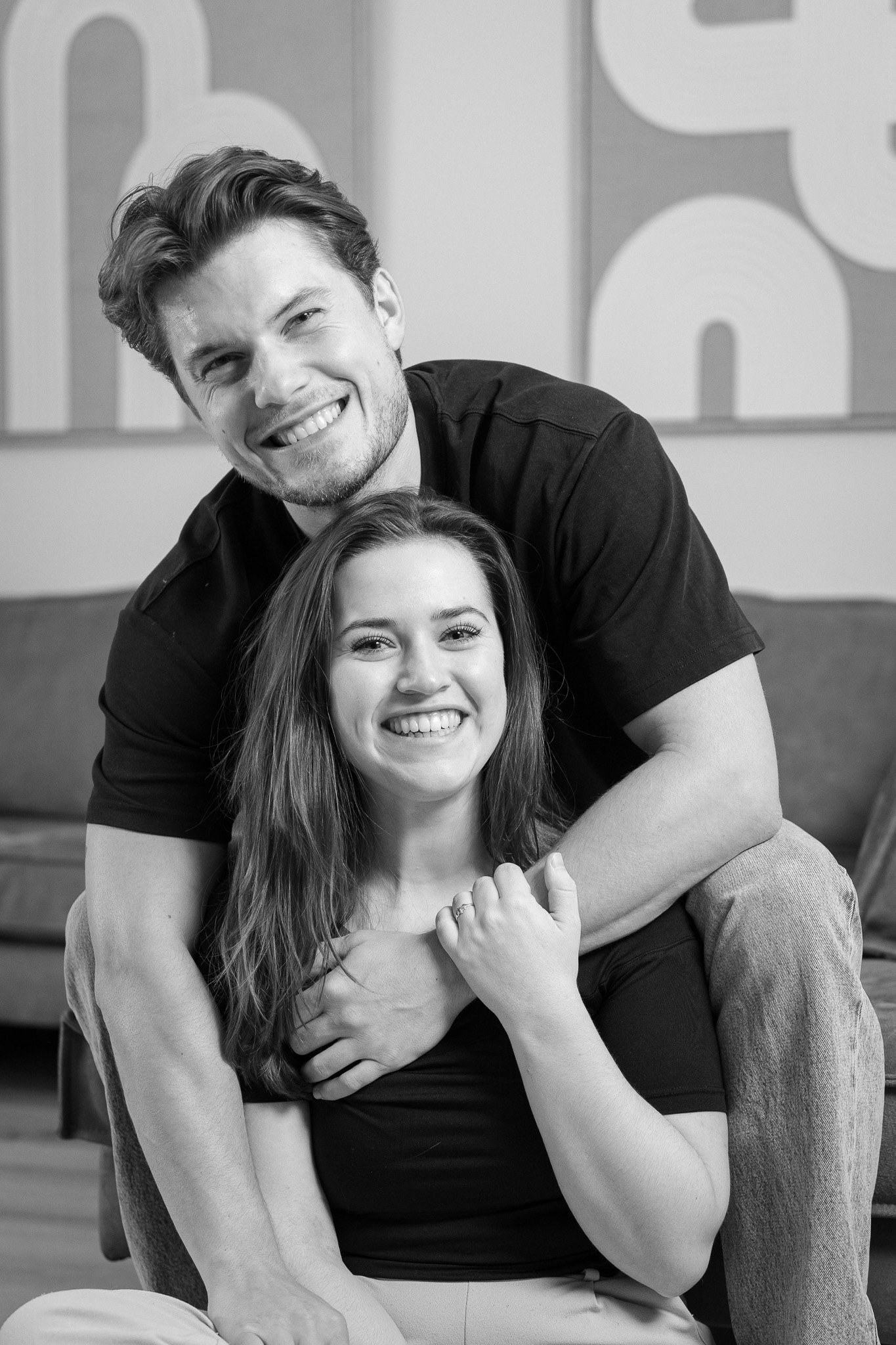 Een jongeman en een jong vrouw die samen lachen, de man staat achter de vrouw en heeft zijn armen om haar heen. De vrouw zit op de grond en de man staat achter haar, beide dragen zwarte shirts. De foto is in zwart-wit en ze zijn in een gezellige kamer met een bank en een kunstwerk op de achtergrond.