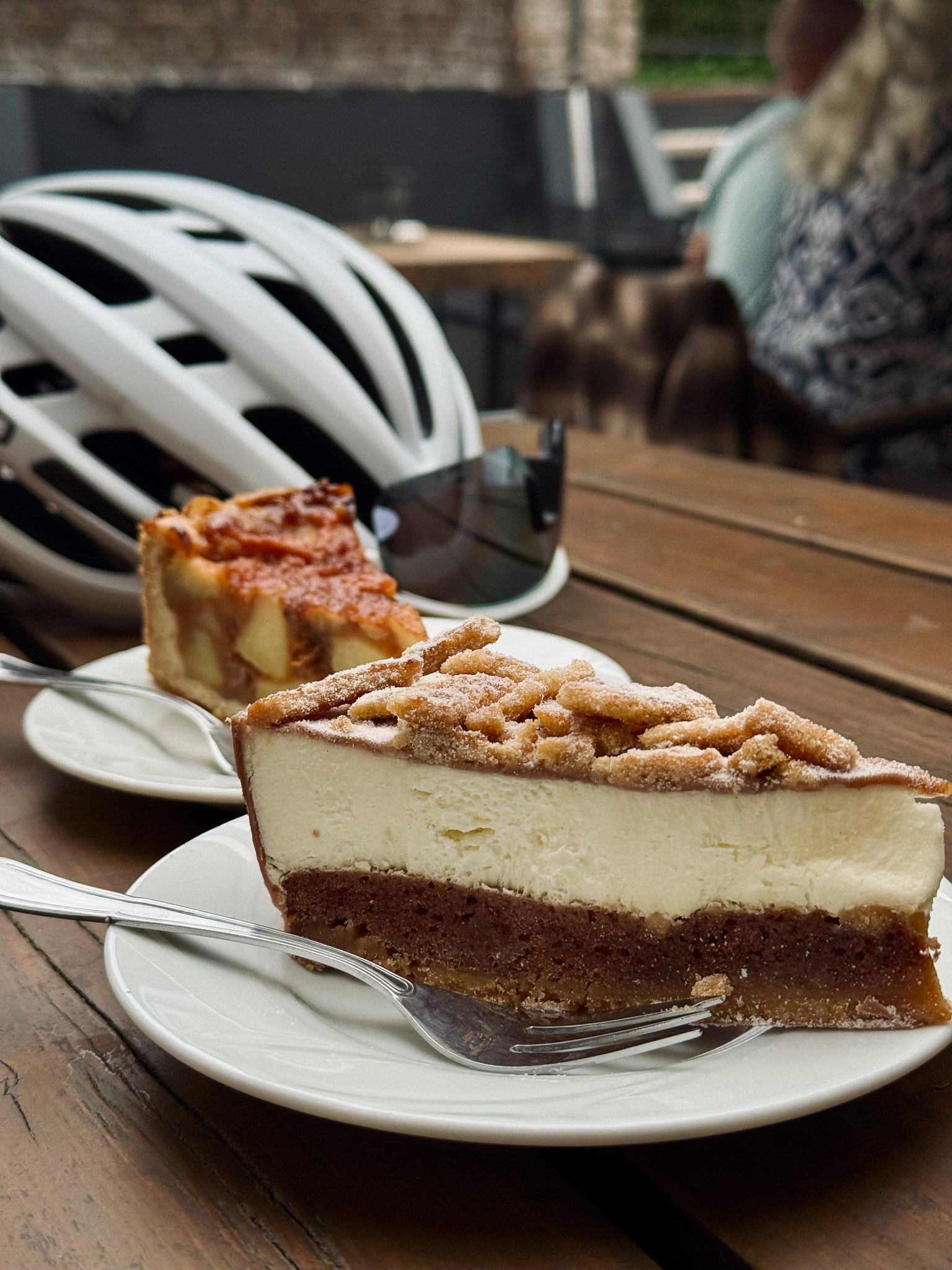 Twee stukken taart op witte borden op een houten tafel, met een fietshelm en een zonnebril op de tafel, op een terras.