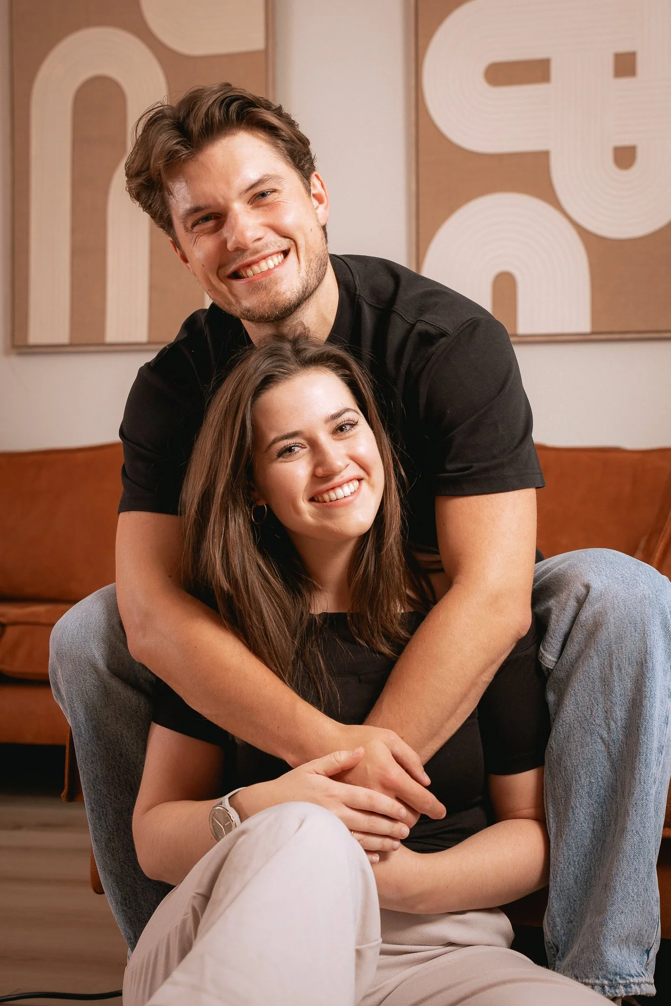 Een jonge man en vrouw poseren samen in een woonkamer, lachend. De man staat achter de vrouw, die op de vloer zit, en houdt haar handen vast.