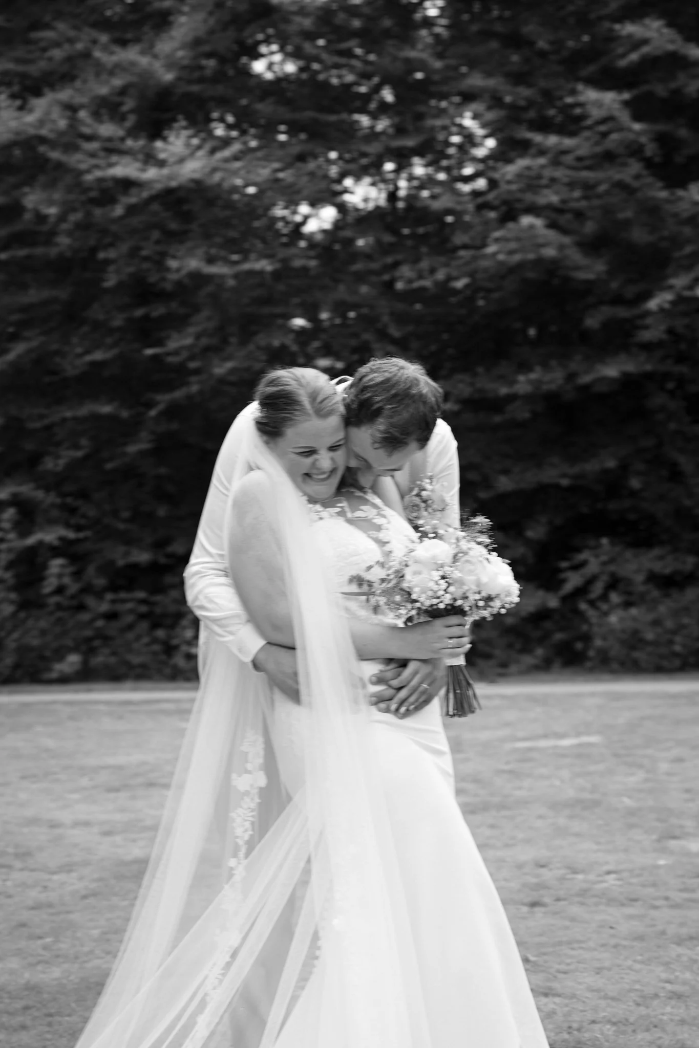 Bruidskoppel in trouwkleding omhelzen en lachen, met een bosrijke achtergrond en een bloemenboeket.