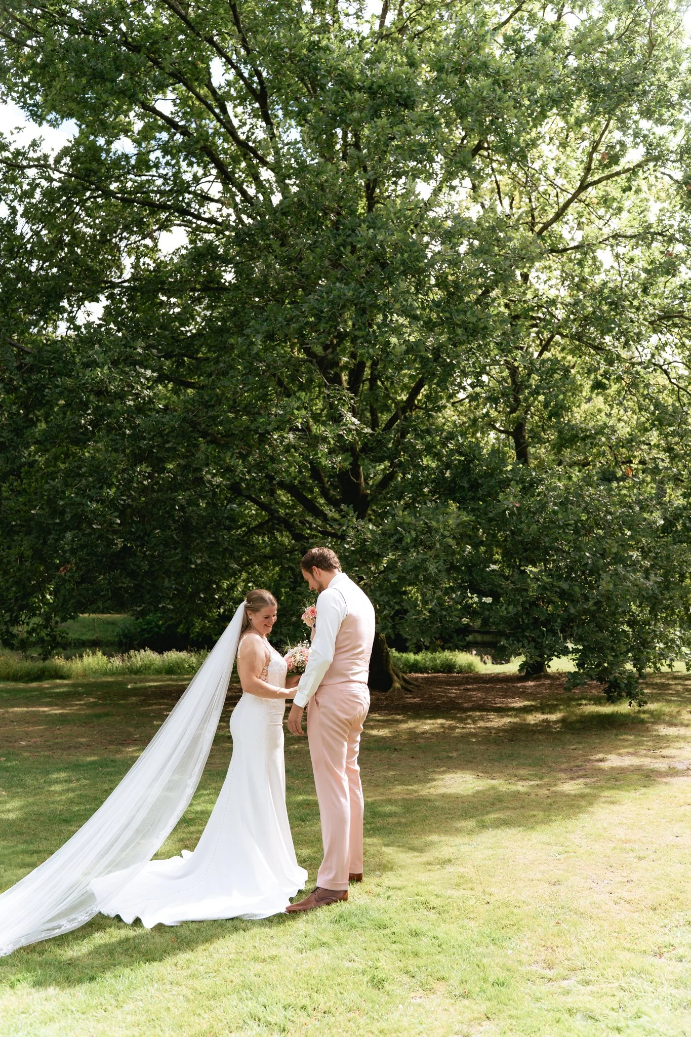 Een bruid en bruidegom staan samen onder een grote groene boom in een groene tuin tijdens hun huwelijk, lieve momenten vastgelegd.