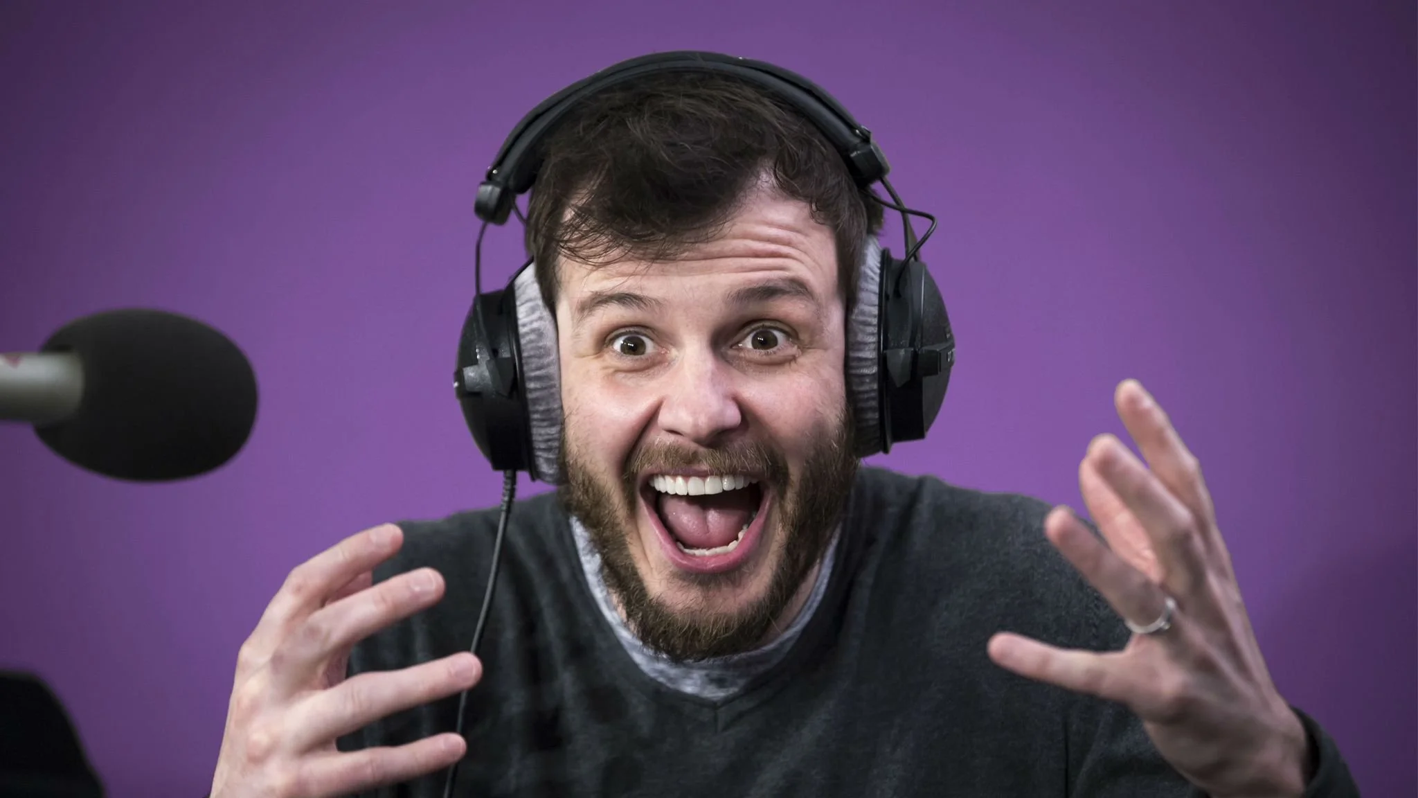 Man with a beard wearing headphones, displaying an excited or surprised expression with mouth open and hands raised, in front of a purple background.