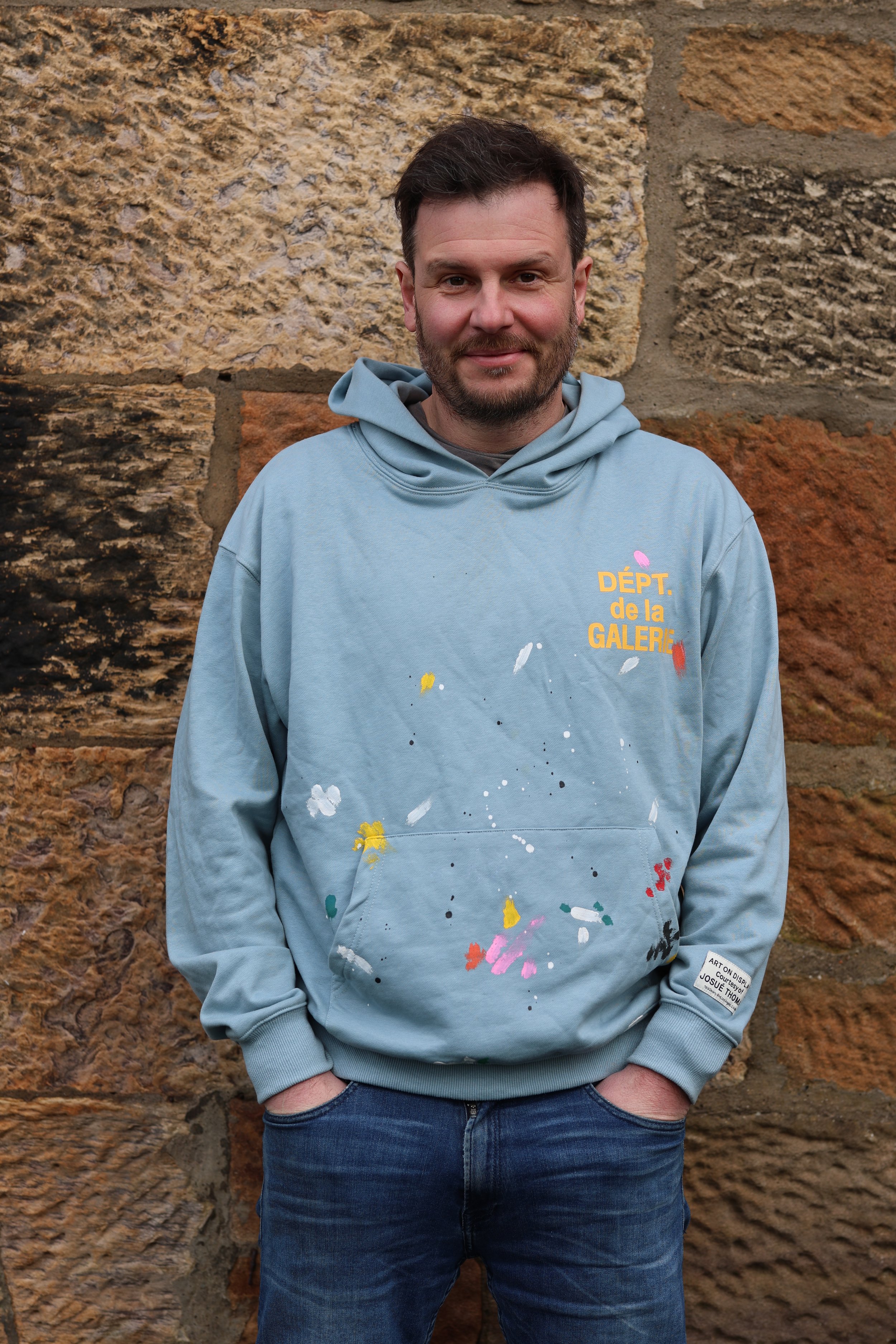 A man with short dark hair and a beard standing against a stone wall, wearing a light blue hoodie with colorful paint splatters and yellow text that reads 'DÉPT. de la GALERIE'.