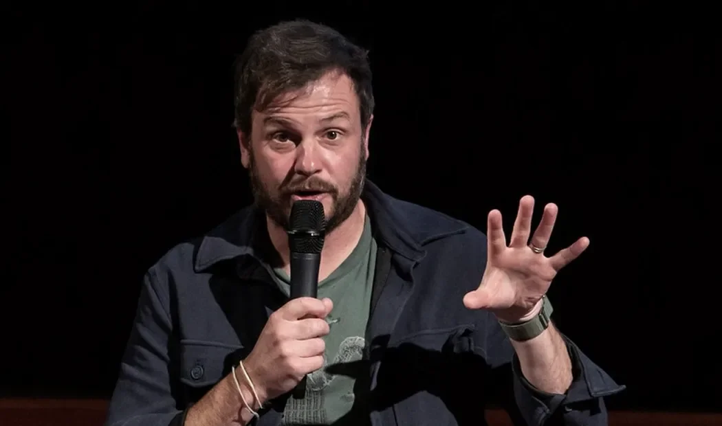 Stand-up comedian performing on stage holding a microphone, gesturing with his left hand, wearing a dark jacket and a green t-shirt, with a dark background.