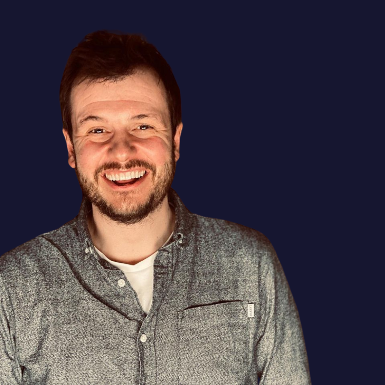 A smiling man with a beard and short brown hair, wearing a gray button-up shirt over a white t-shirt, standing against a dark background.
