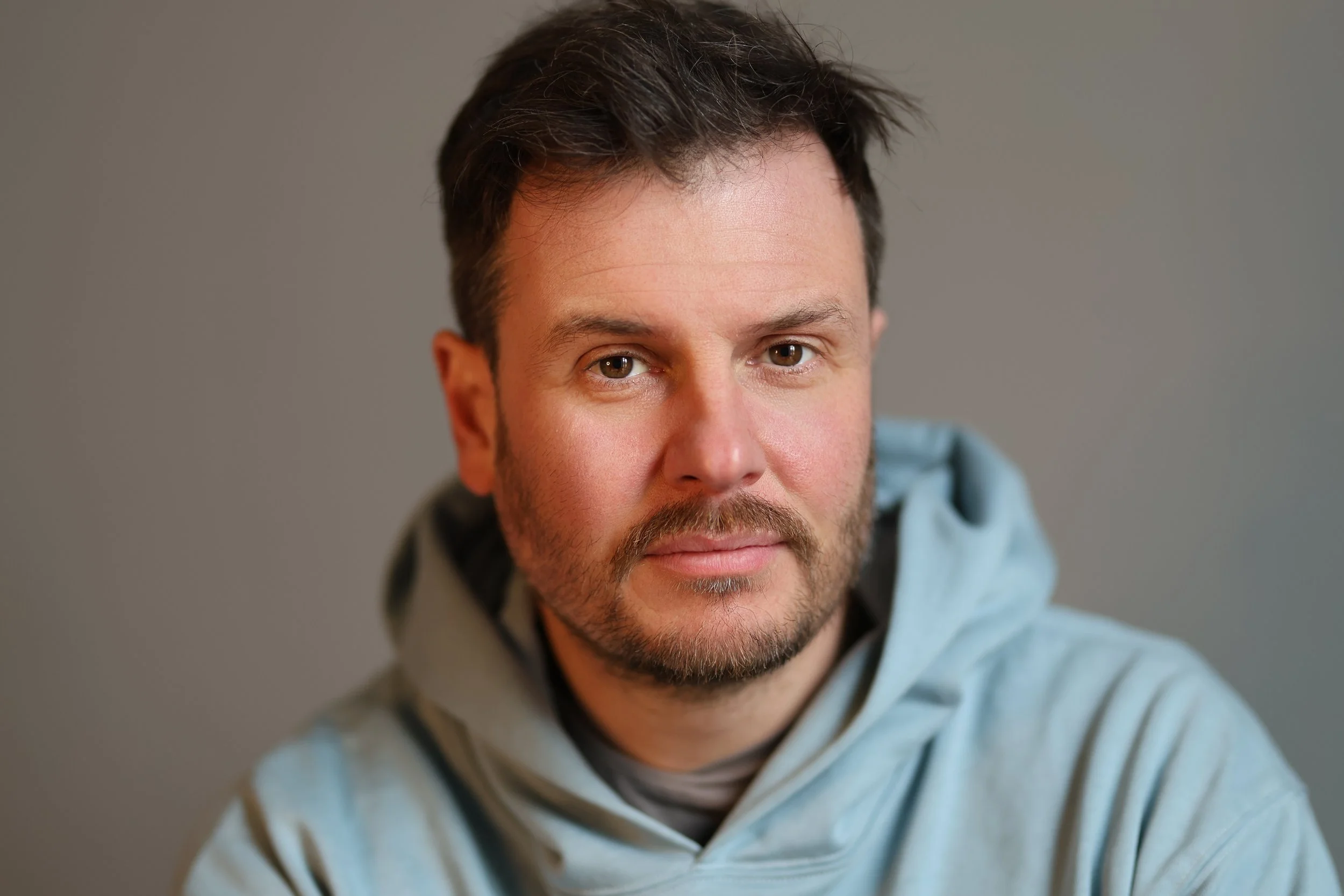 A man with brown hair, brown eyes, and a beard, wearing a light blue hoodie, looking directly at the camera with a neutral expression.