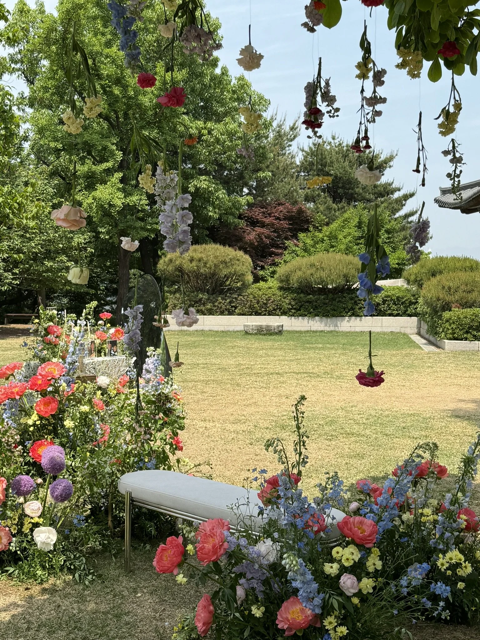 A garden scene with hanging and potted flowers, green trees, and a small grassy area, with a single white bench in the foreground.