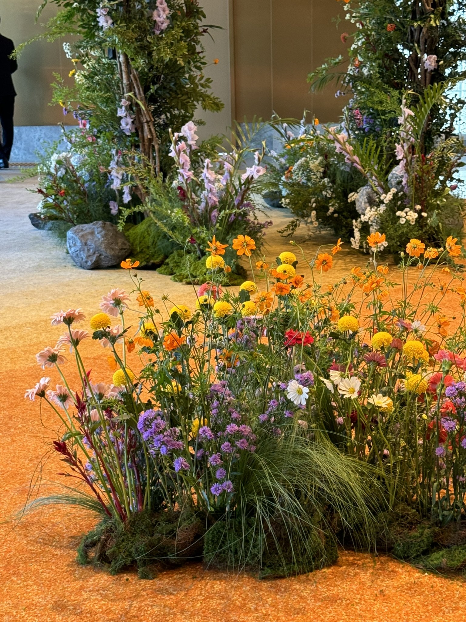 Colorful flower arrangements with pink, yellow, orange, purple, and white flowers placed on an orange carpeted floor, with some greenery and rocks in the background.