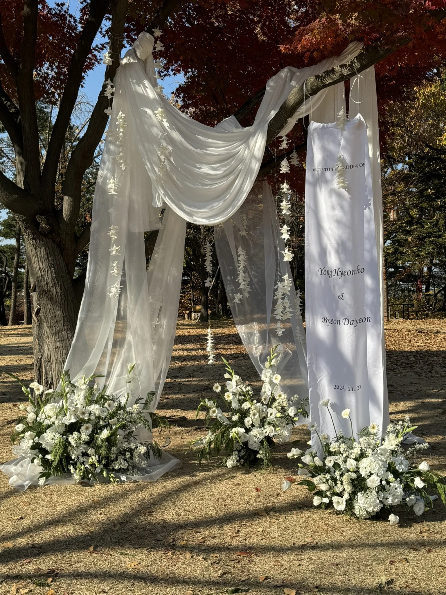 Wedding arch draped with white fabric and decorated with hanging white leaves, set outdoors on a grassy area with trees and fallen leaves in the background. Floral arrangements of white flowers are placed at the base of the arch.