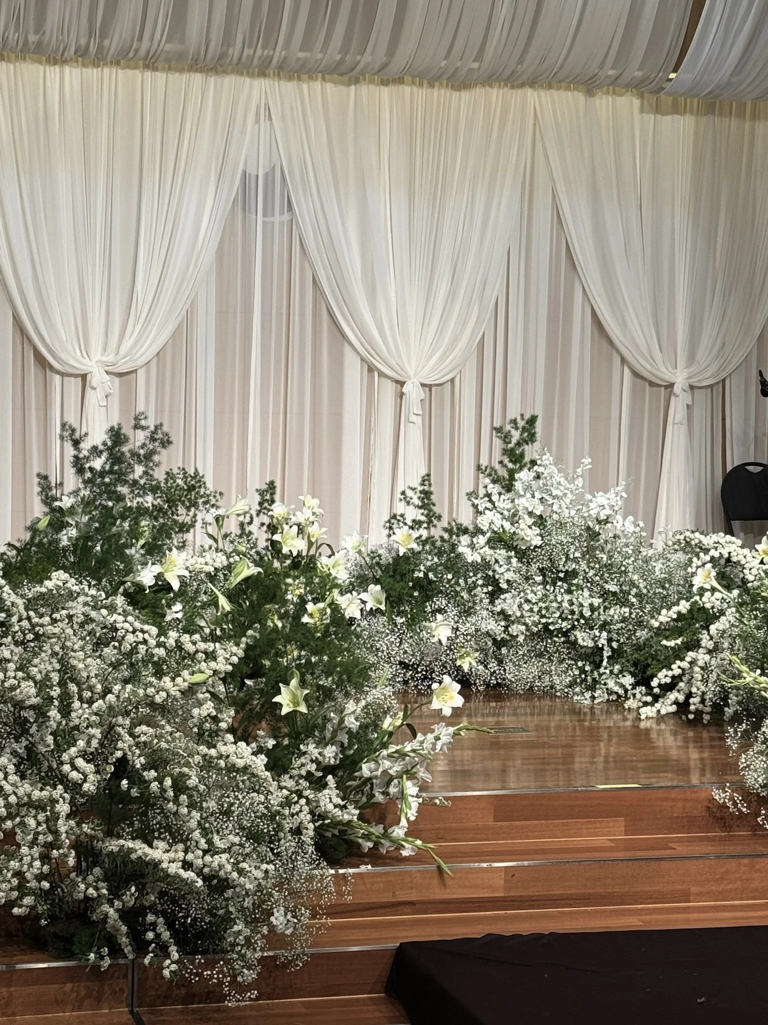 White draped curtains with floral arrangements of white lilies, baby's breath, and greenery at a wedding altar.