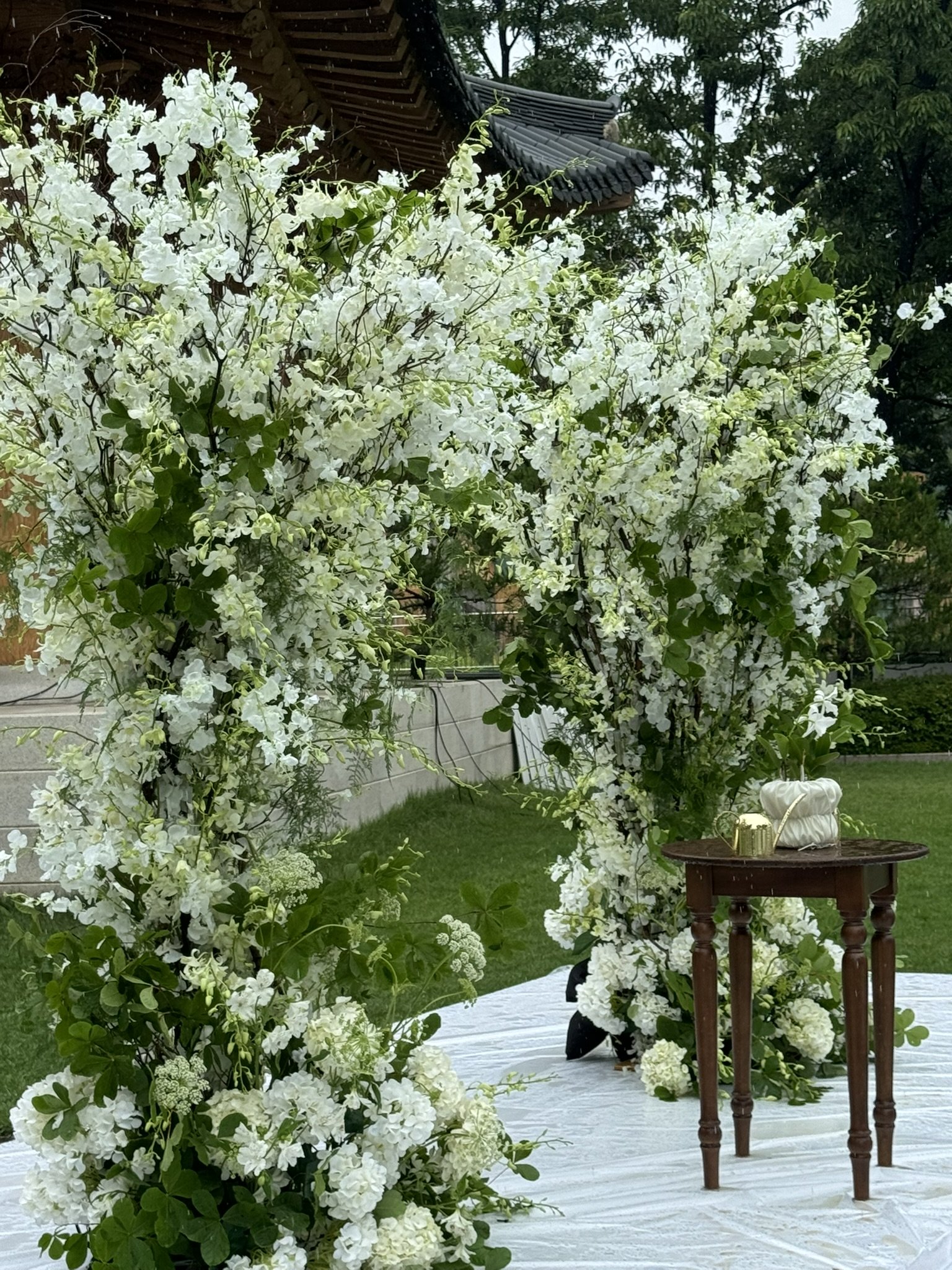 A floral arch made of white blossoms and green leaves, decorated for an outdoor wedding or event, with a small wooden table holding a white cloth, gold pitcher, and small items. The setting is grassy, with a building with traditional Asian roof tiles