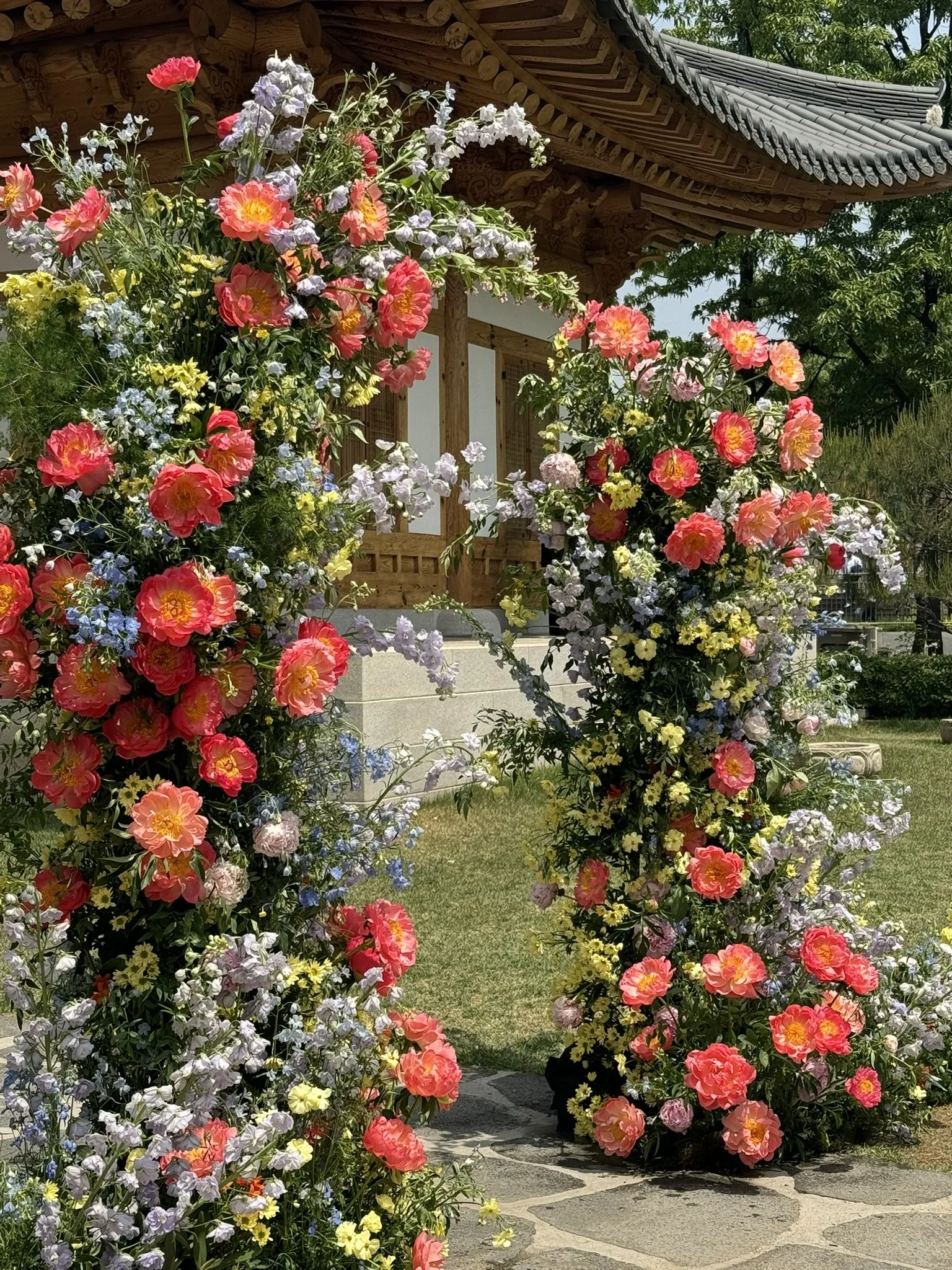 A floral archway with pink, purple, and yellow flowers in front of a traditional wooden Asian building with a curved tiled roof.