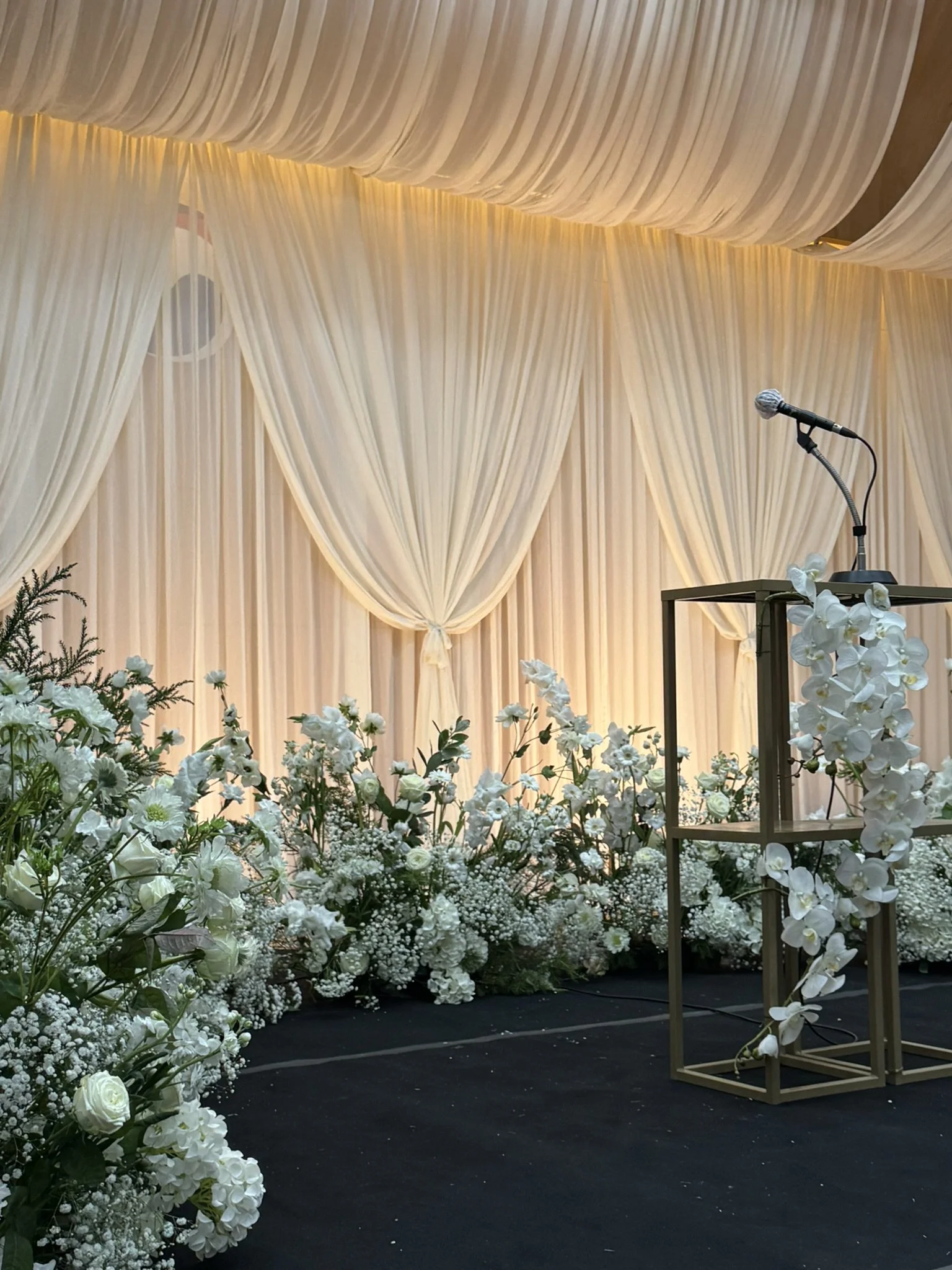 A decorated stage with beige draped curtains, surrounded by white flowers and greenery, with a microphone stand on a wooden podium.