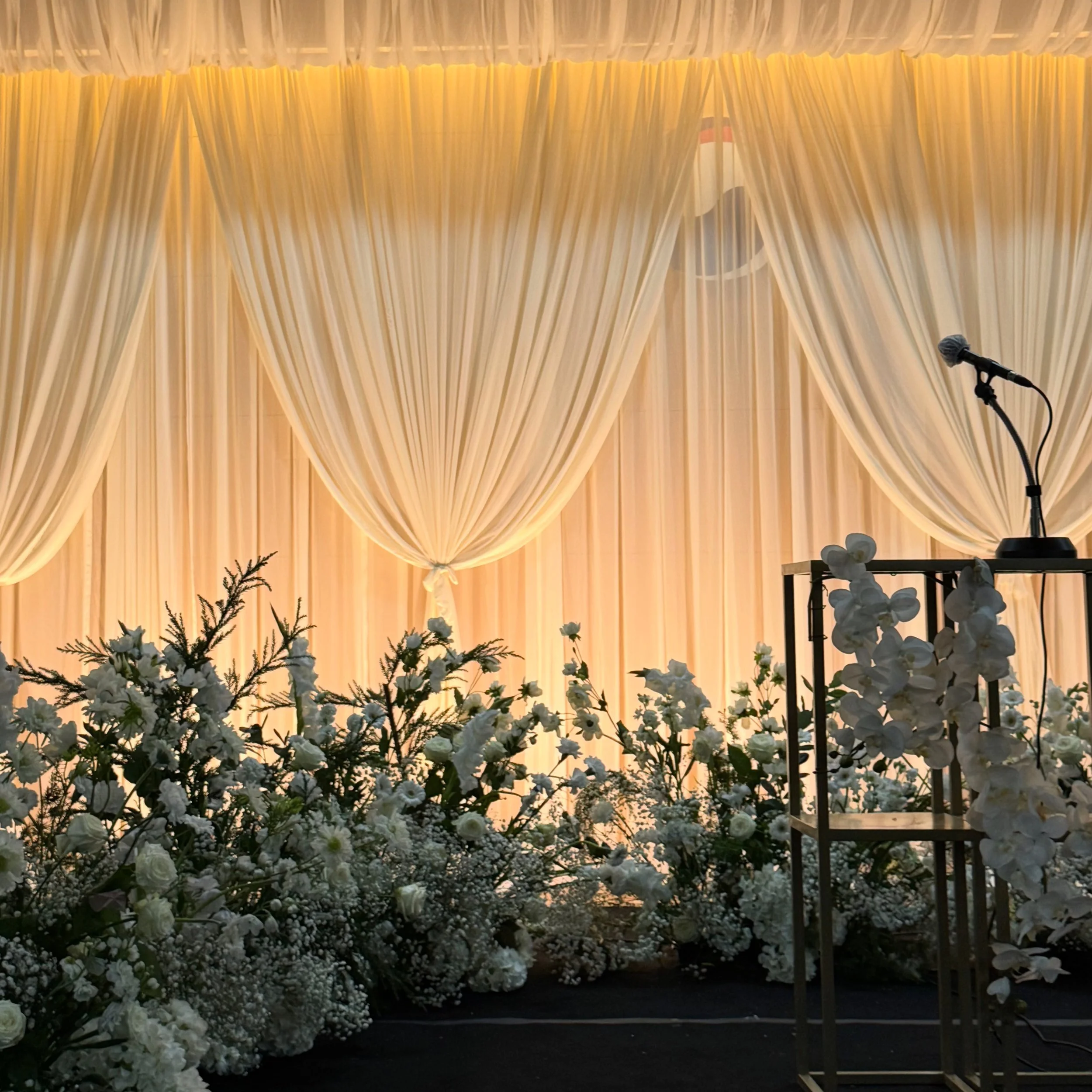 Elegant event stage with cream-colored drapes, floral arrangements of white flowers and greenery, and a lectern with a microphone.