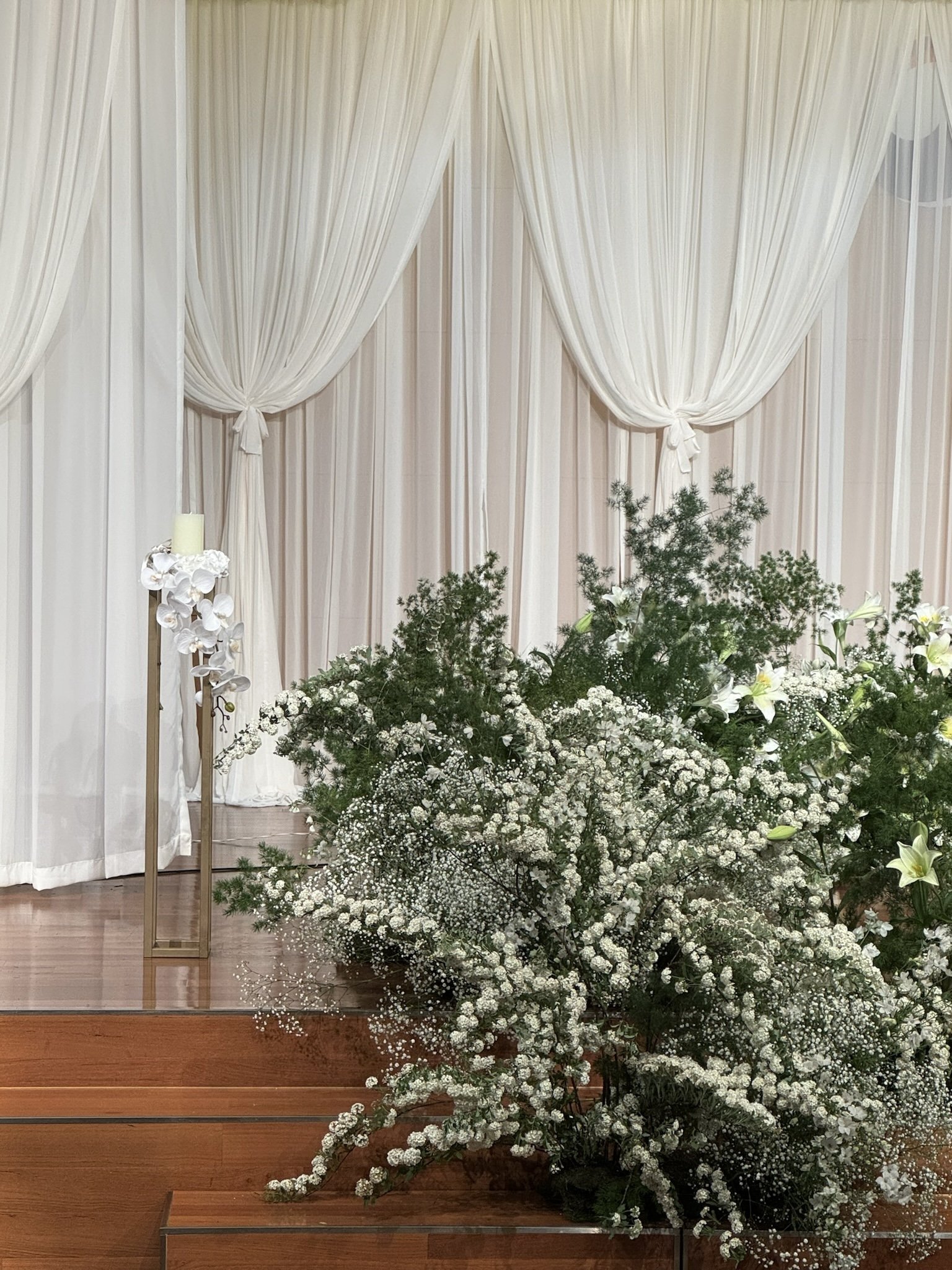 Elegant altar with white curtains, floral arrangements of white baby's breath, roses, and lilies on a wooden stage.
