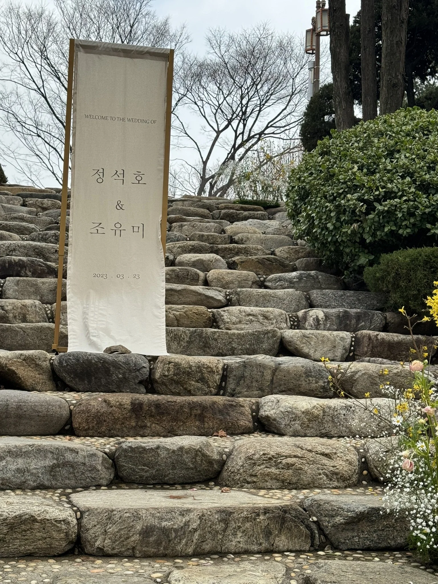 A stone staircase outdoors leading up to a wedding event, with a welcome banner in Korean and English, trees with no leaves in the background, and some greenery and flowers on the side.