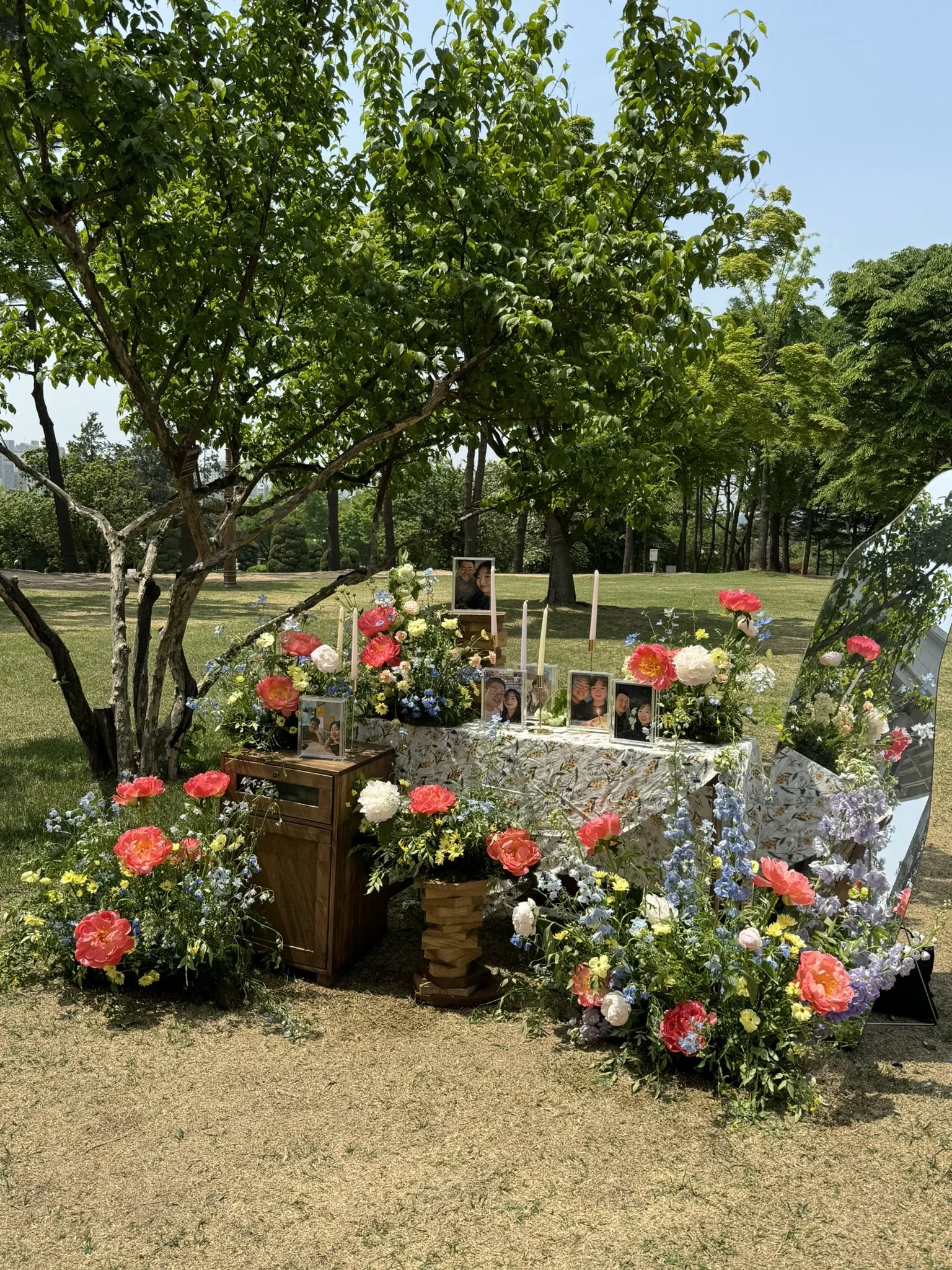Outdoor memorial setup with framed photos, candles, and floral arrangements under a tree in a park.