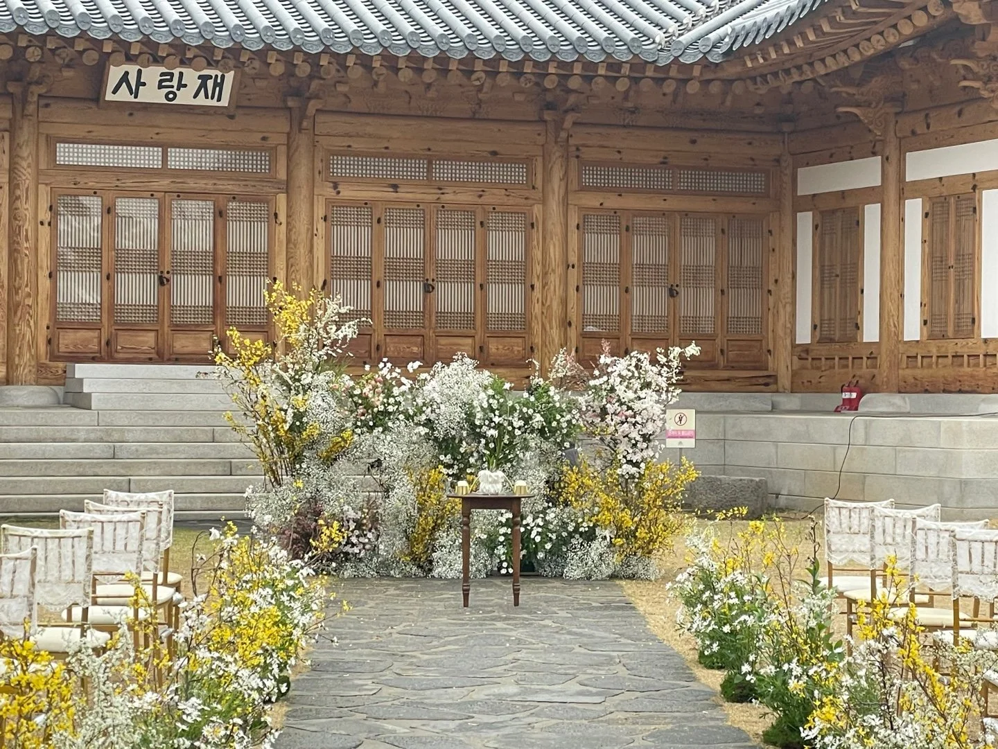 Traditional Korean wedding venue with a wooden building, stone steps, and floral decorations including white and yellow flowers along a stone pathway.