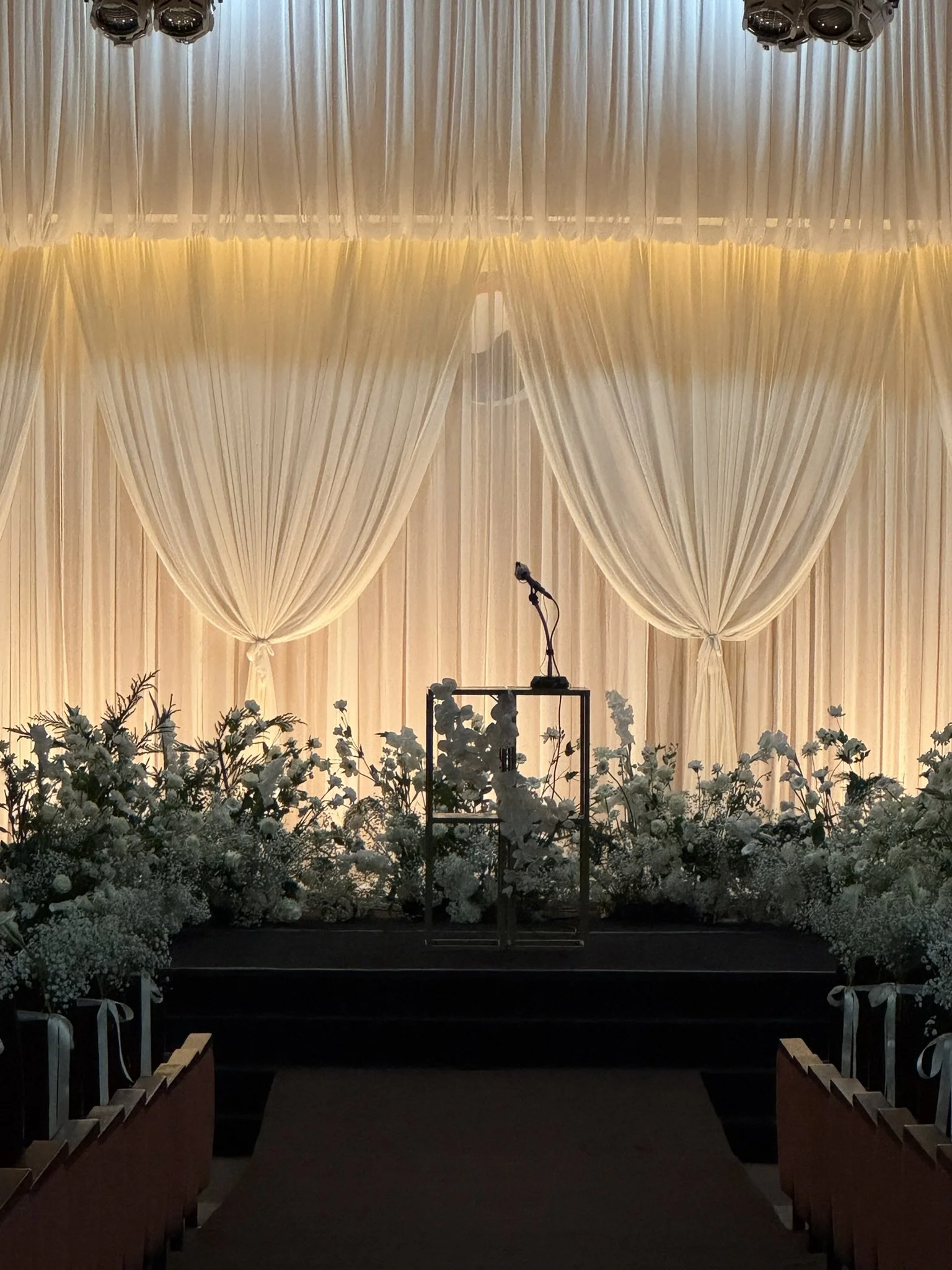 Empty stage with a microphone on a stand, decorated with white flowers and draped beige curtains in the background, in a decorated event hall.