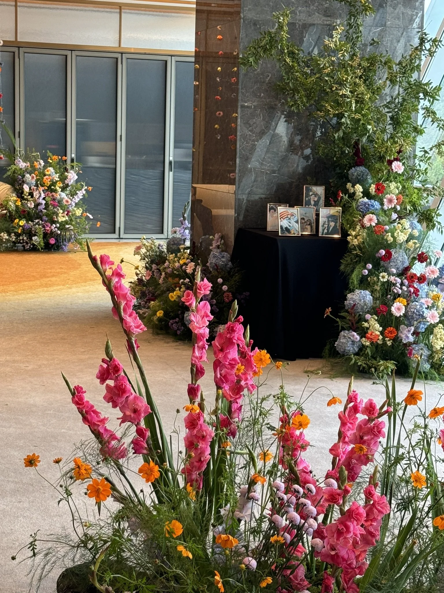 Elegant interior space with a floral display including pink gladiolus and orange flowers, a black table with framed photos, and abundant greenery and additional flowers in the background.