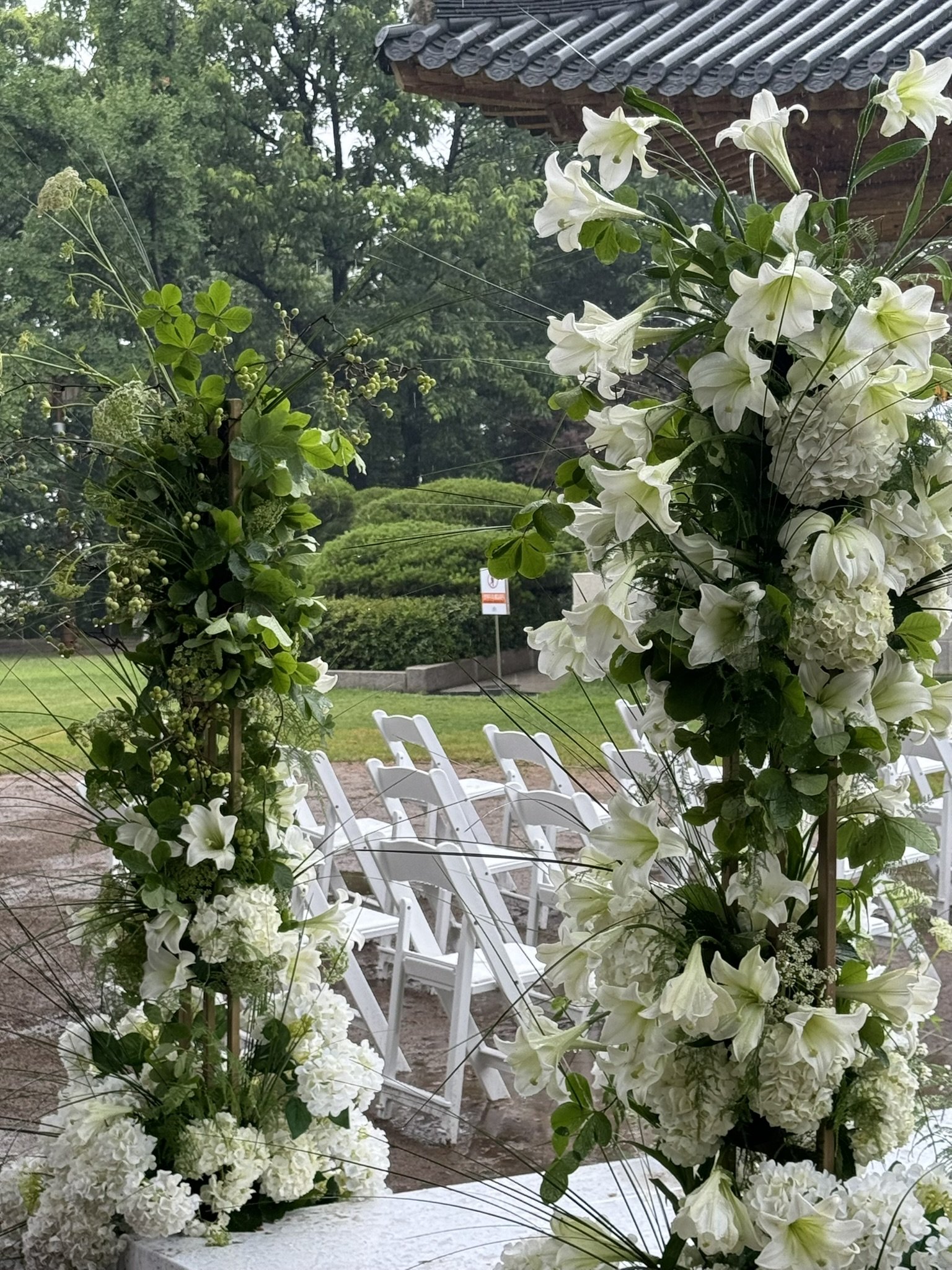 Close-up of a floral arrangement of white lilies, hydrangeas, and greenery, with white chairs arranged outdoors in the background, likely set up for a wedding ceremony.