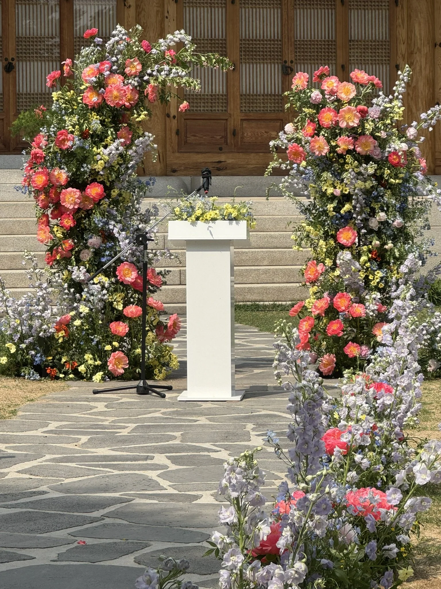A decorated outdoor stage with a white podium, surrounded by colorful flowers, set against a wooden building with steps.