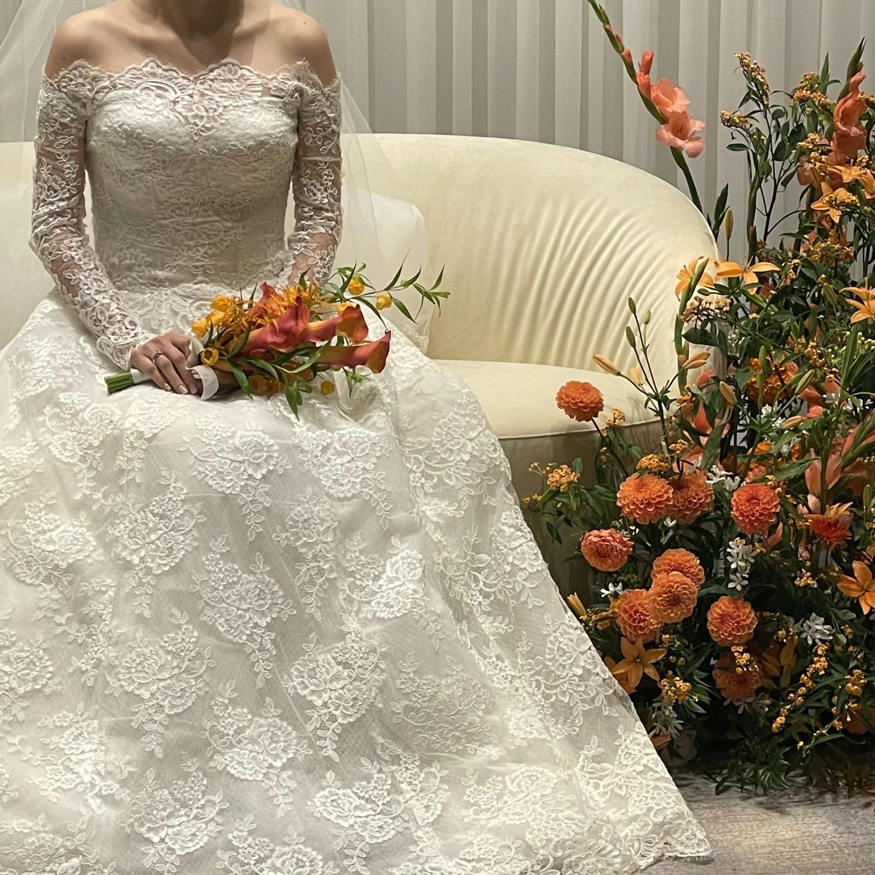 A bride sitting on a cream-colored sofa, wearing a long-sleeved lace wedding dress, holding a bouquet of orange, pink, and yellow flowers, with a large floral arrangement beside her.