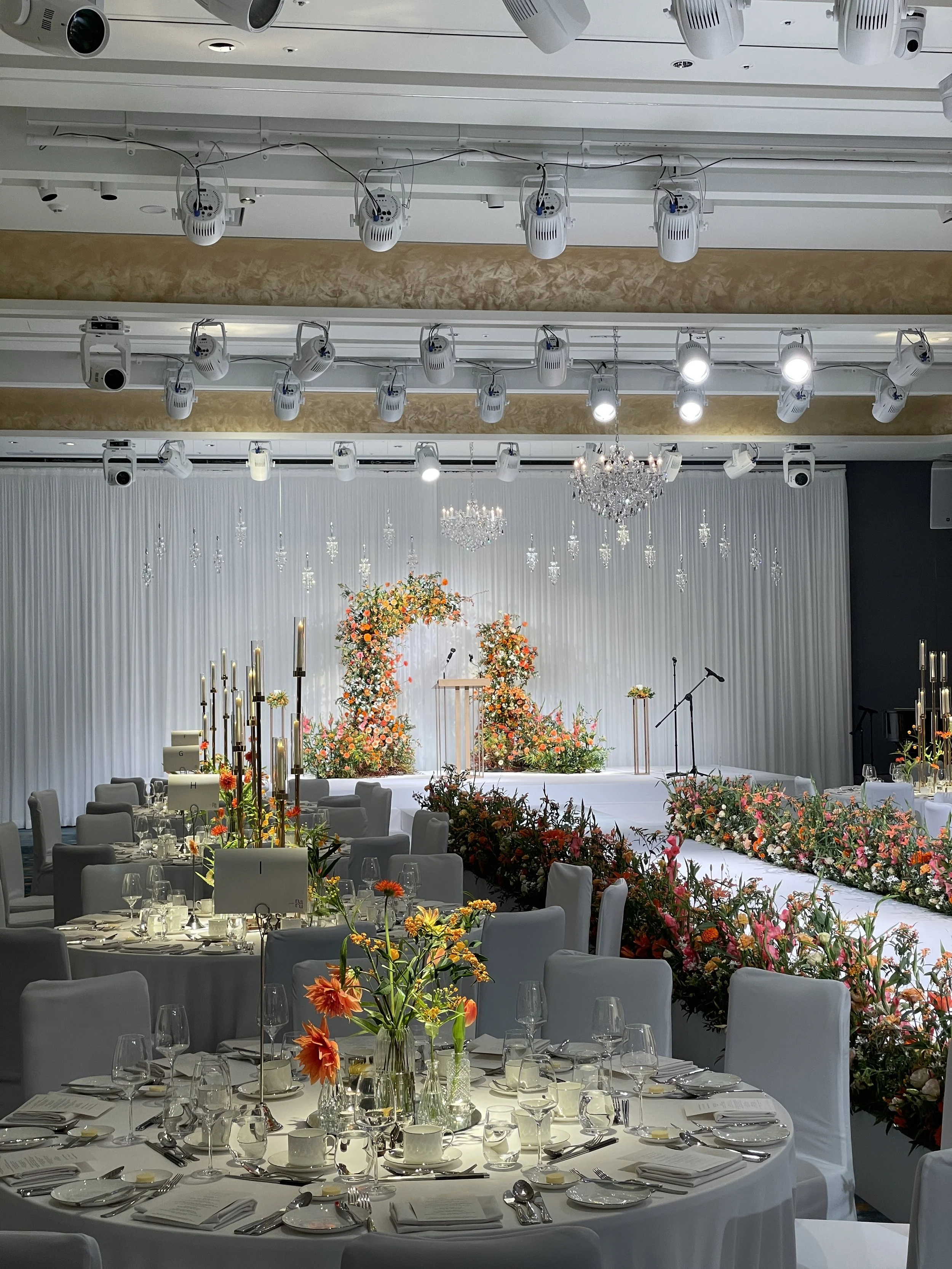 Elegant banquet hall decorated for a wedding or event with round tables set with glassware and floral centerpieces, a stage with floral arrangements and podium, chandeliers, and professional ceiling lighting.