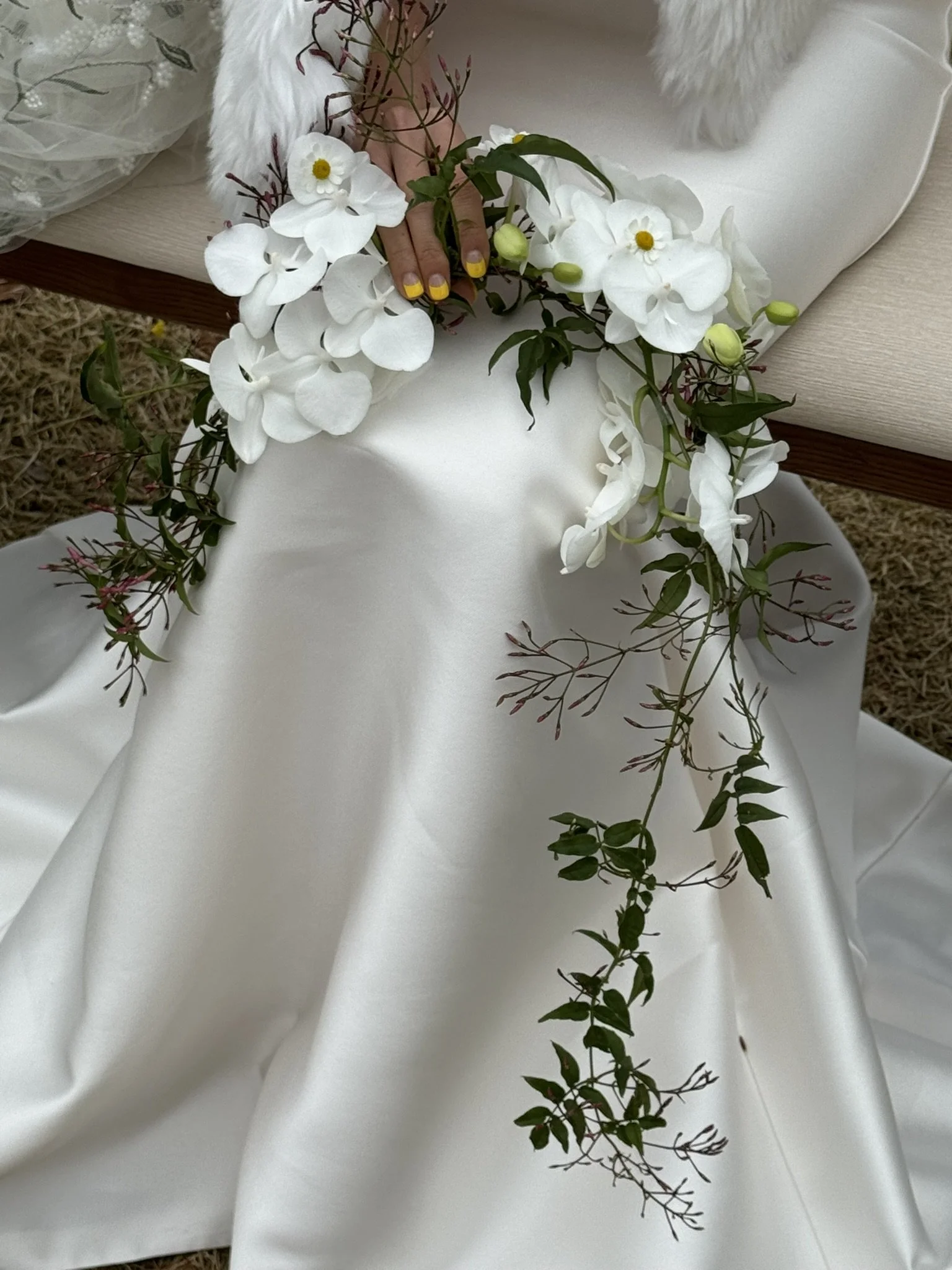 Person in a white dress holding a floral crown made of white orchids and greenery, seated on a white surface outdoors.