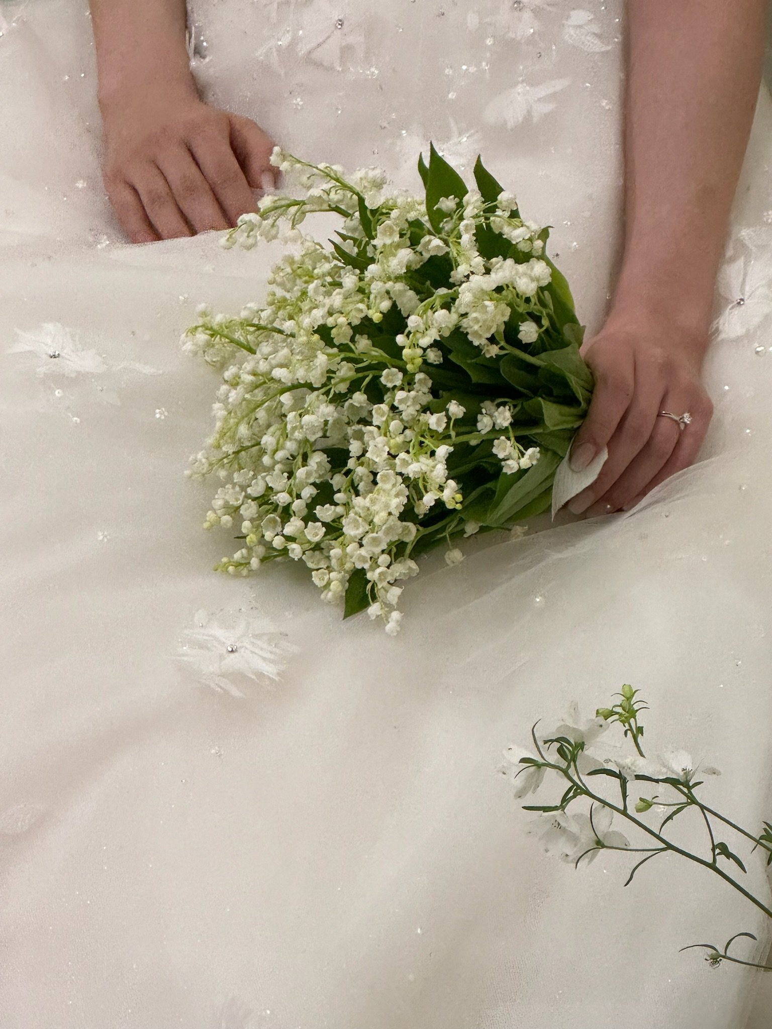 A person in a wedding dress holding a bouquet of white flowers, with a small branch of similar flowers lying on a white fabric surface.