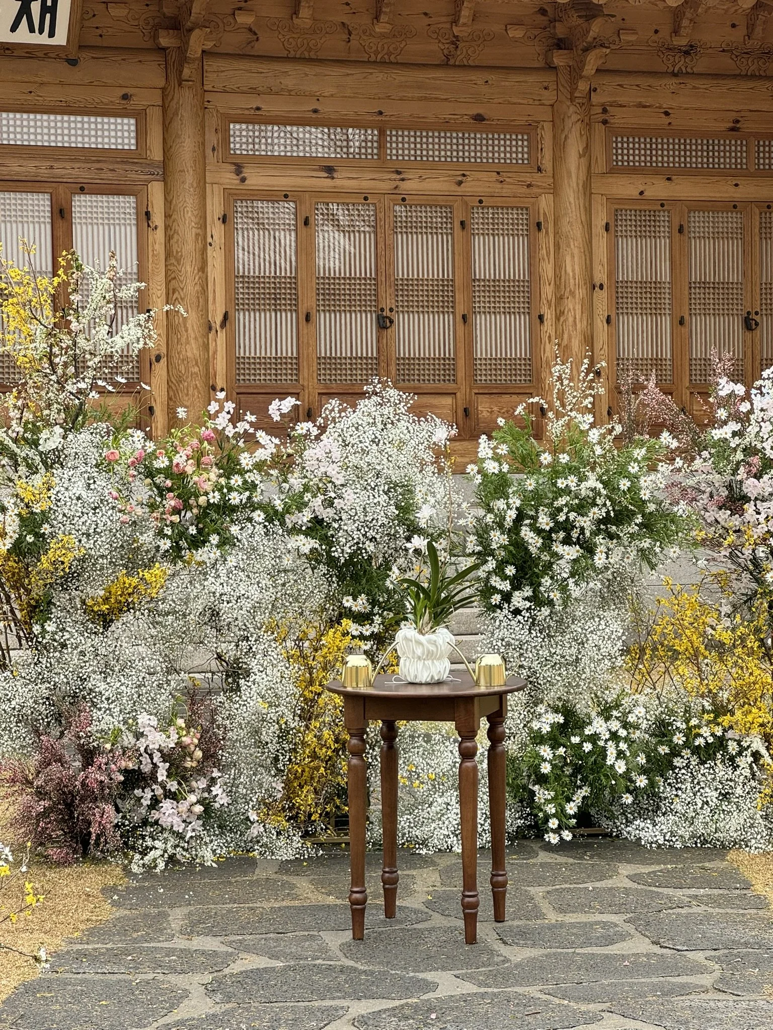 A small wooden table with a potted plant and two golden cups on top, surrounded by white, pink, yellow, and purple flowers against a wooden lattice wall.
