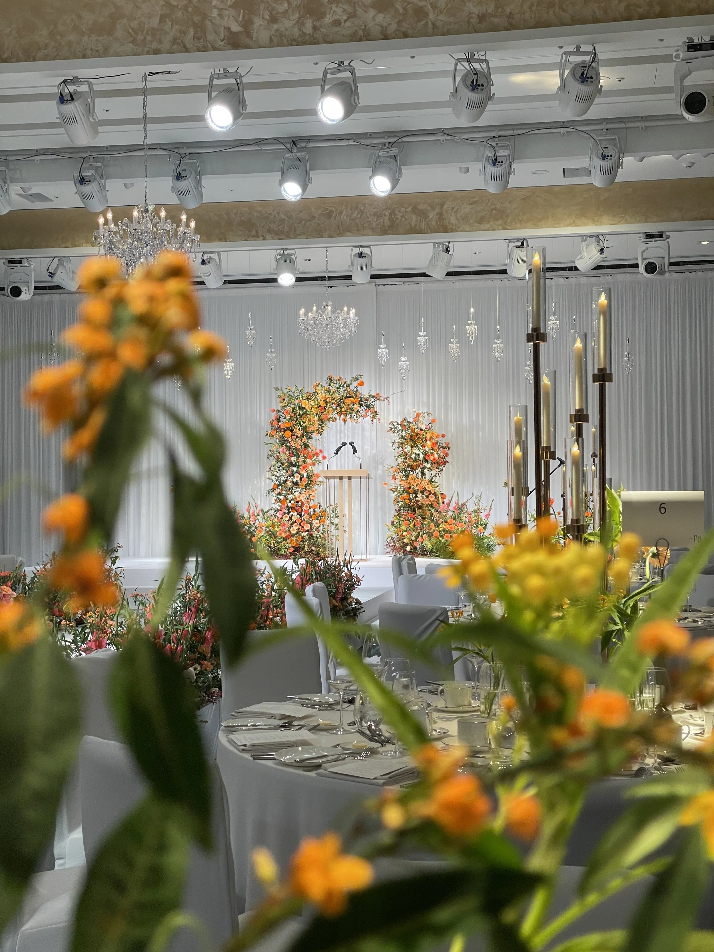 Decorated event banquet hall with round tables, floral arrangements, chandeliers, and stage with podium.