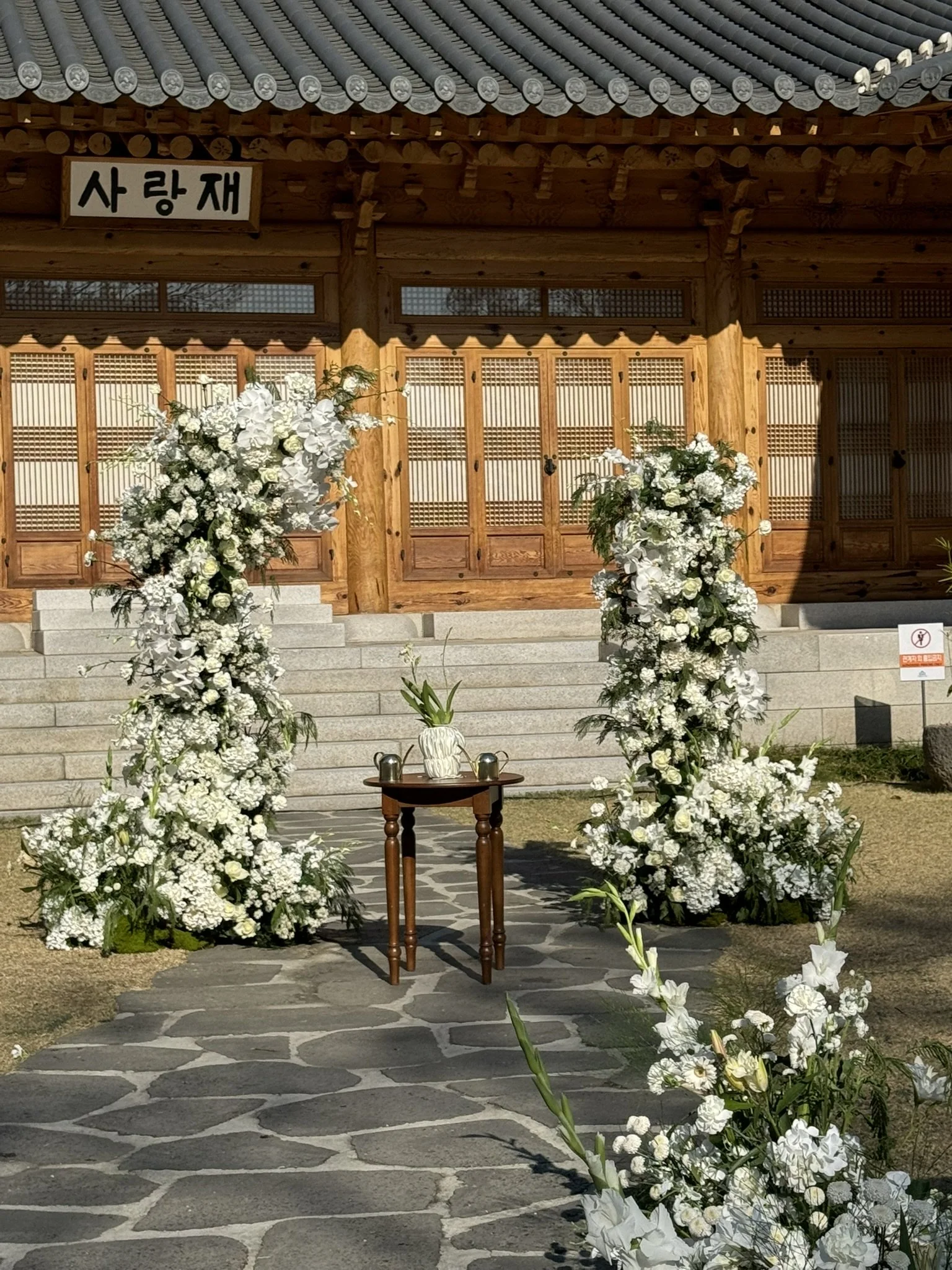 Decorated wedding altar with white flowers, a small wooden table with a white vase and flowers, set in front of a traditional wooden building with Korean characters on a sign.