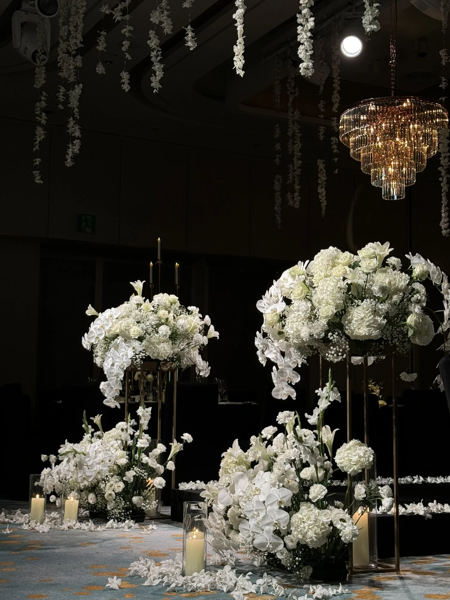 Elegant wedding or event decor featuring large arrangements of white flowers including roses, hydrangeas, and orchids, with tall candles and a gold chandelier hanging from the ceiling in a dimly lit room.
