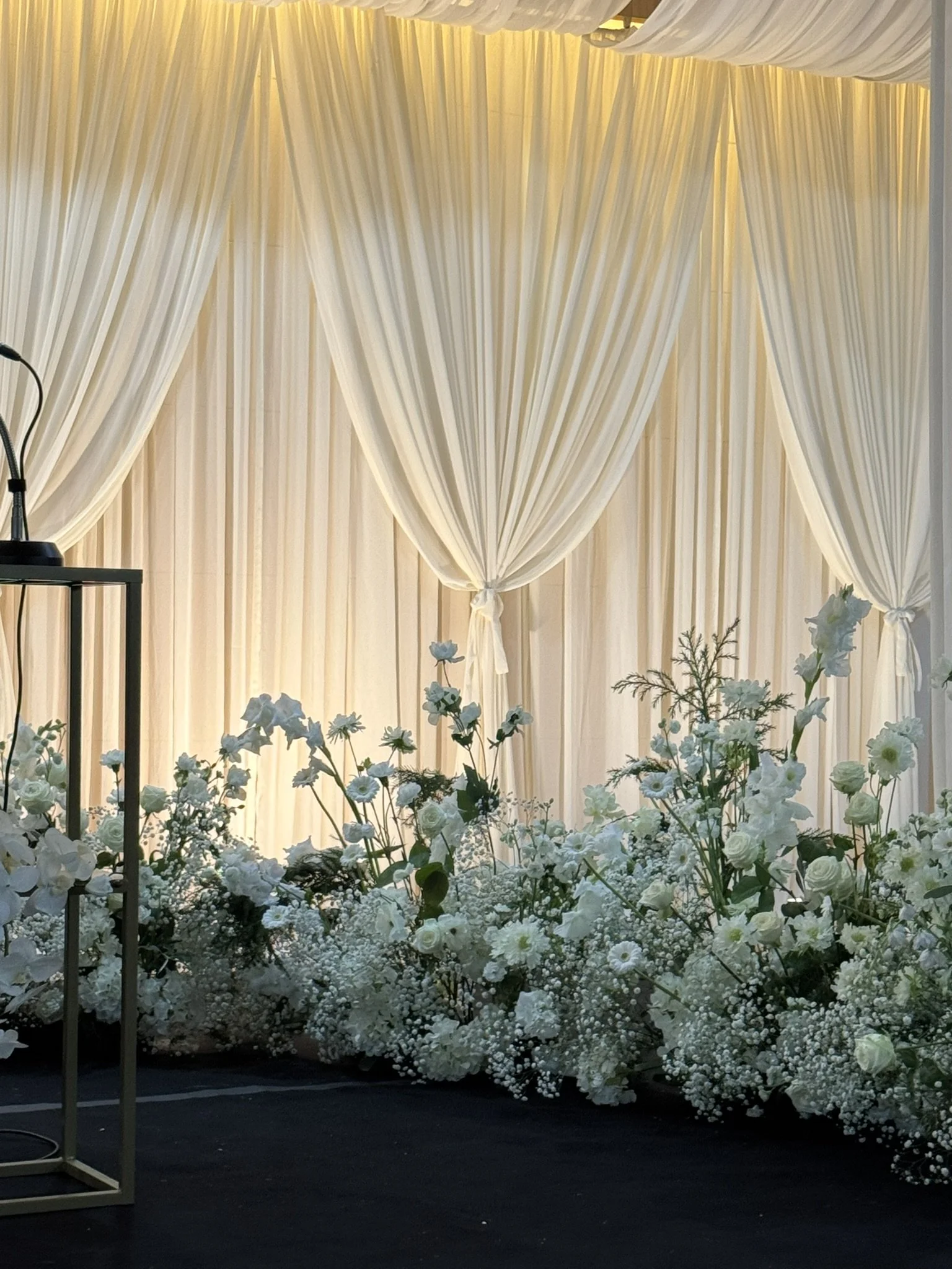 A wedding altar with cream-colored drapes tied back in the center, behind a line of white floral arrangements on the floor, with a microphone stand and a small black table on the left.