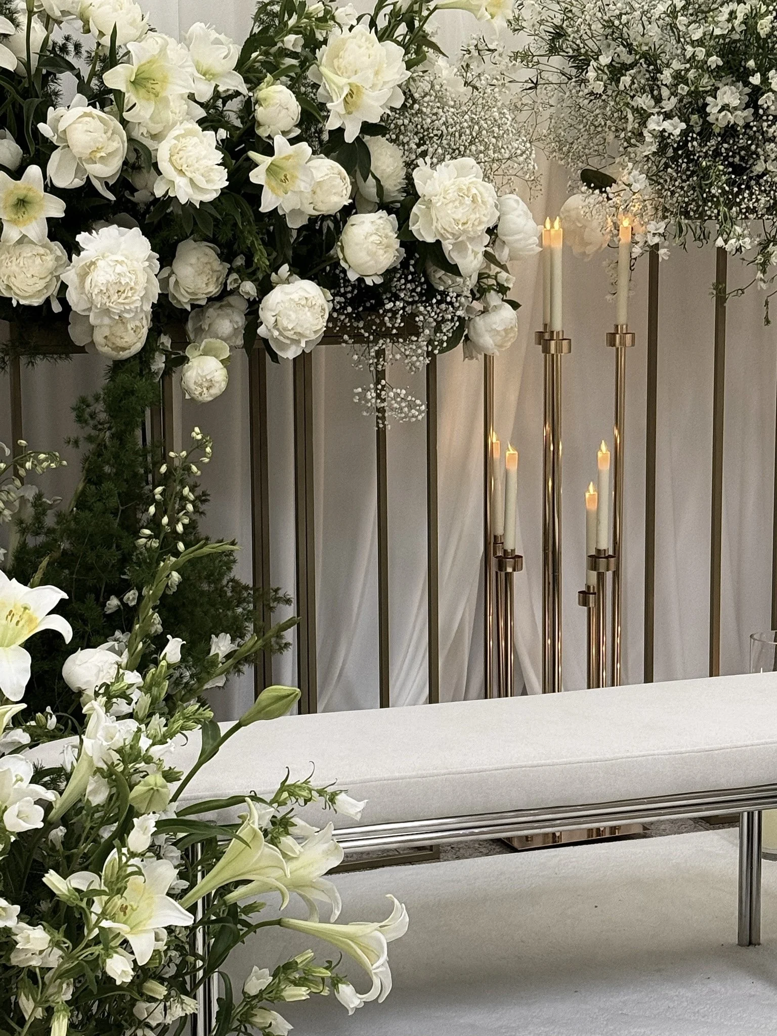 Elegant floral arrangement with white peonies, roses, and lilies, alongside tall white candles on gold stands, set against a white backdrop.