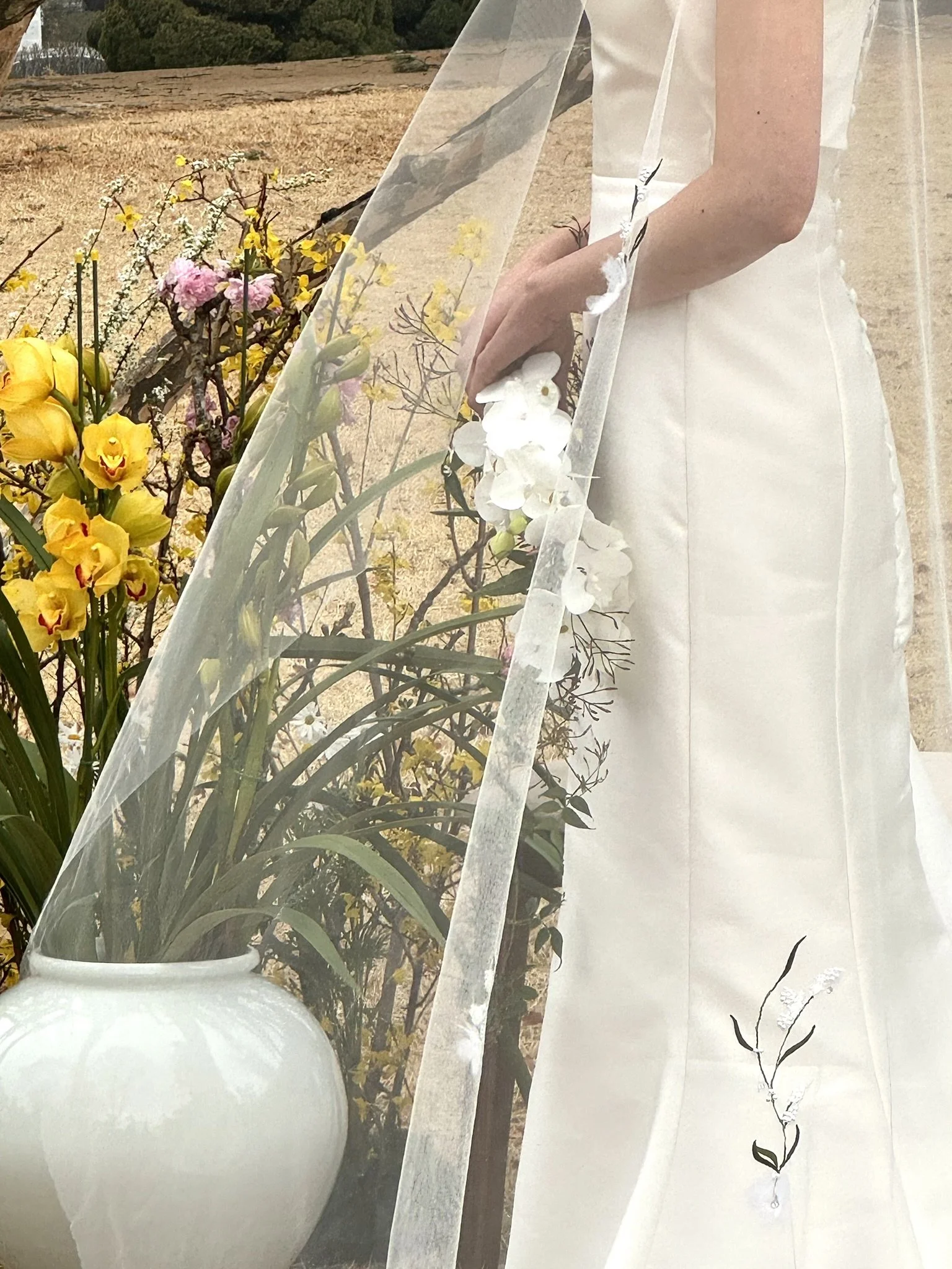 Close-up of a woman wearing a white dress and veil, holding a bouquet of white flowers, standing next to a large white vase with yellow and pink flowers outdoors.