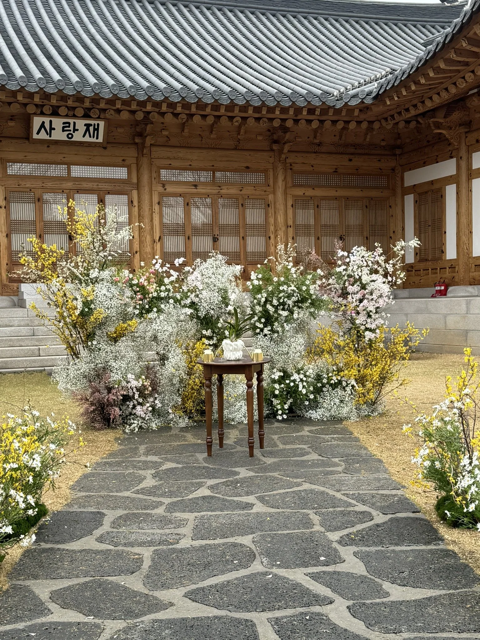 Flower arrangement in front of a traditional wooden Korean building with a tiled roof, with a small table holding a plant and gold containers in the center.