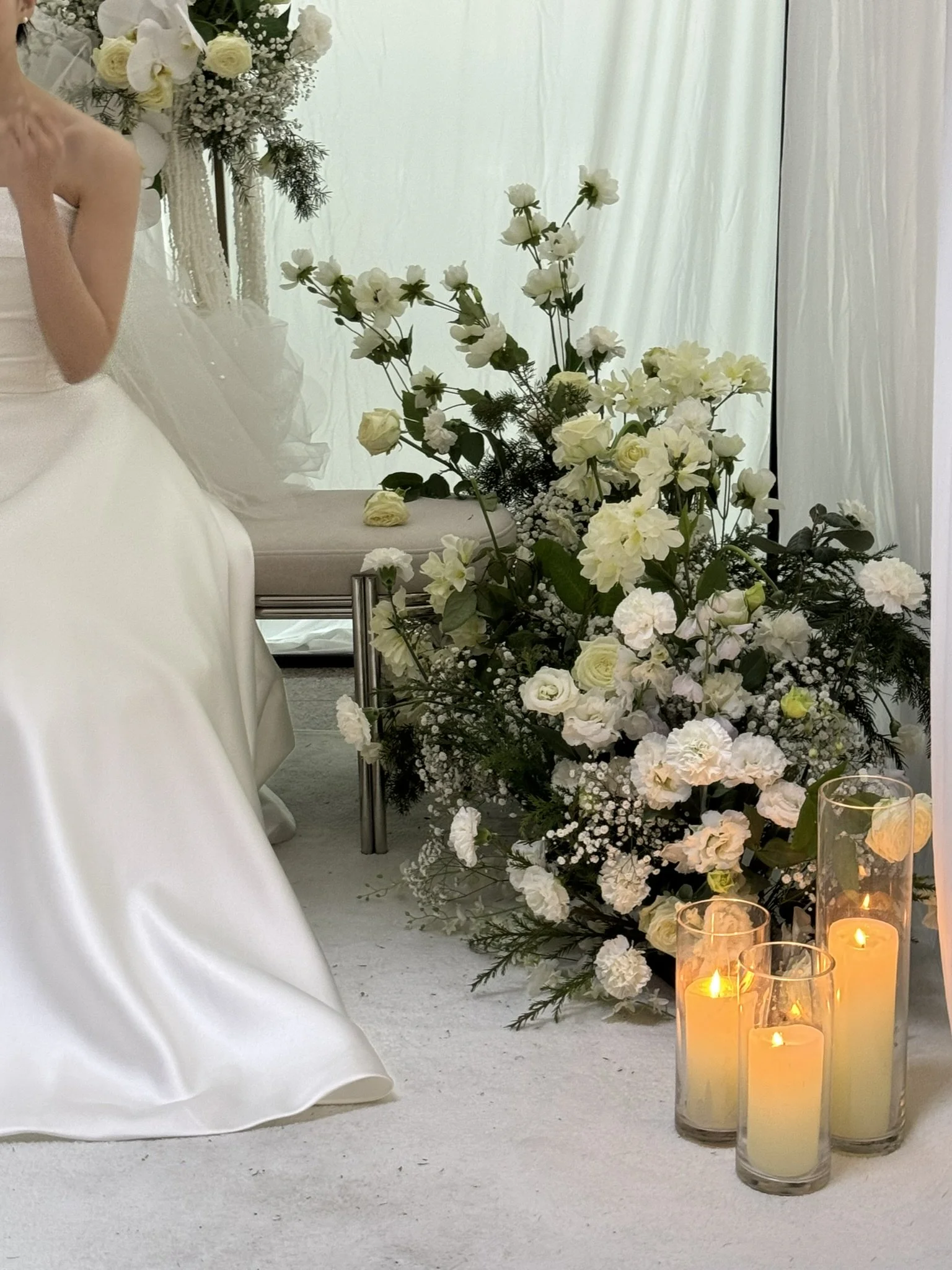 A large floral arrangement of white flowers with candles in glass holders on the floor at a wedding ceremony. A bride in a white wedding dress is partially visible on the left side.