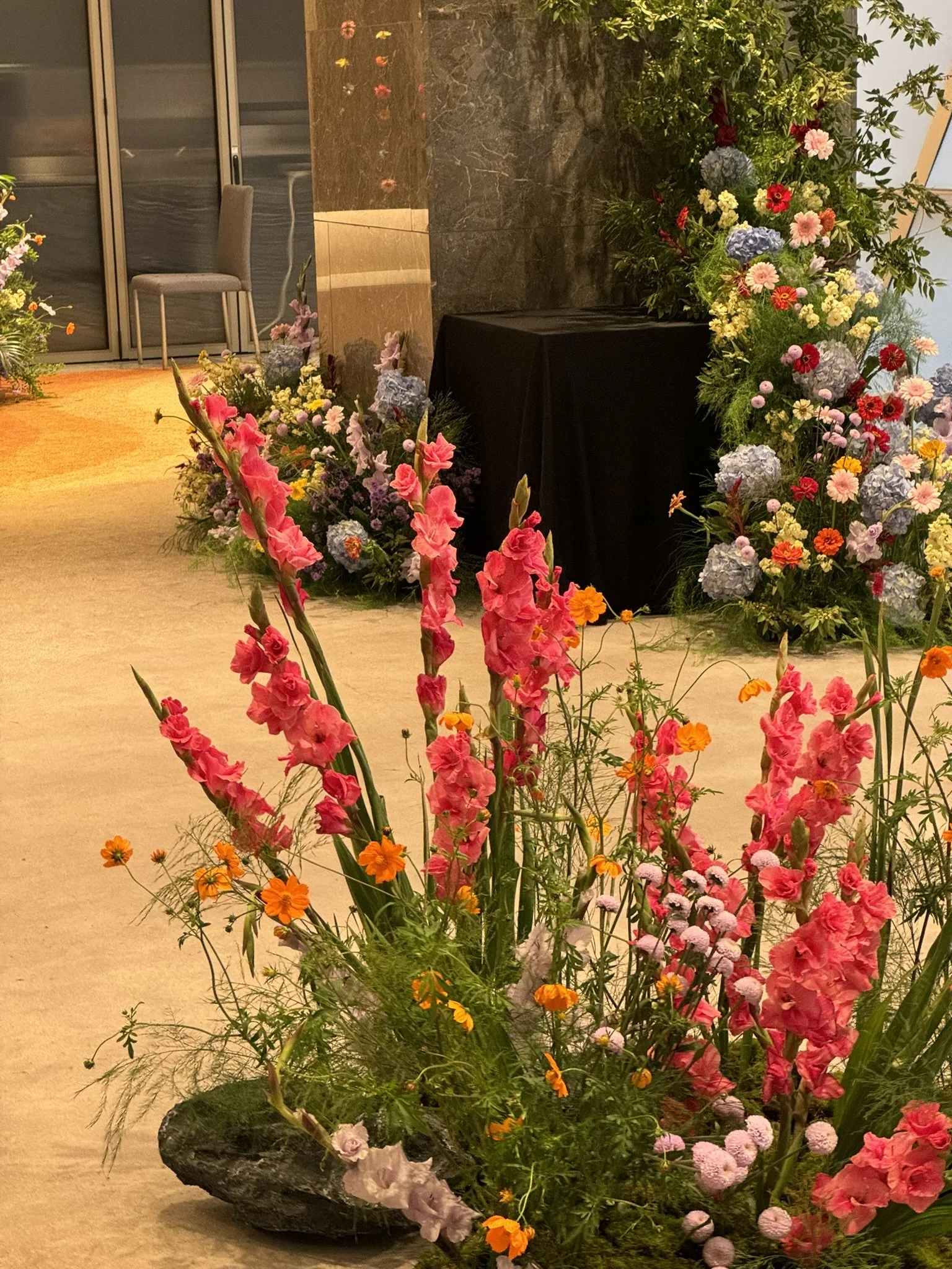 Indoor floral arrangement with pink, purple, red, and orange flowers in a large rock container, set against a backdrop of various other flowers and plants, with a beige carpeted floor.